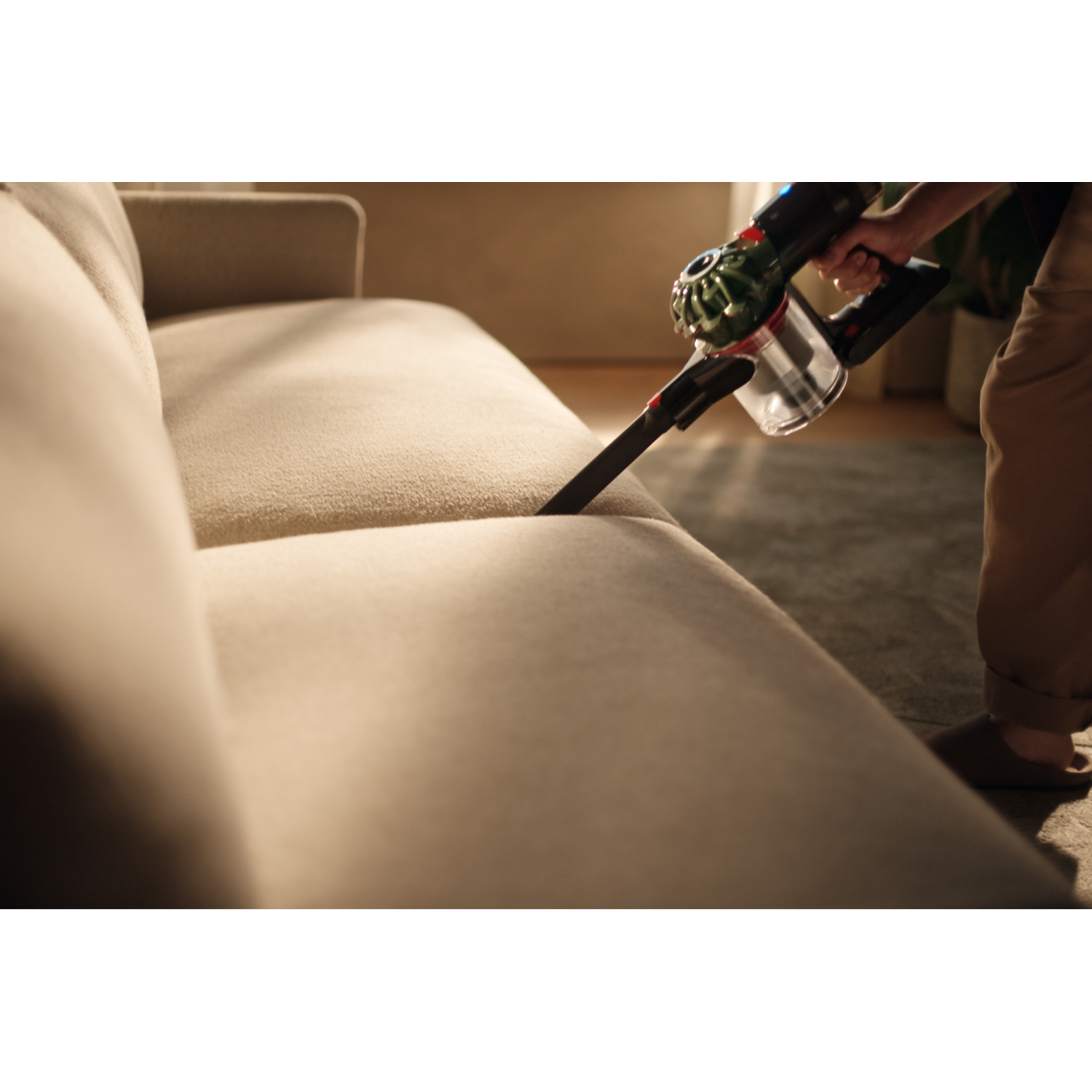 A person vacuums a beige sofa with a handheld vacuum cleaner. The vacuum is black and green. The floor is visible.