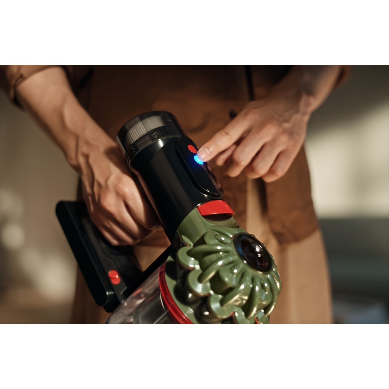 A person's hands operate a green and black cordless vacuum. The person presses a button with a blue light. Brown shirt.