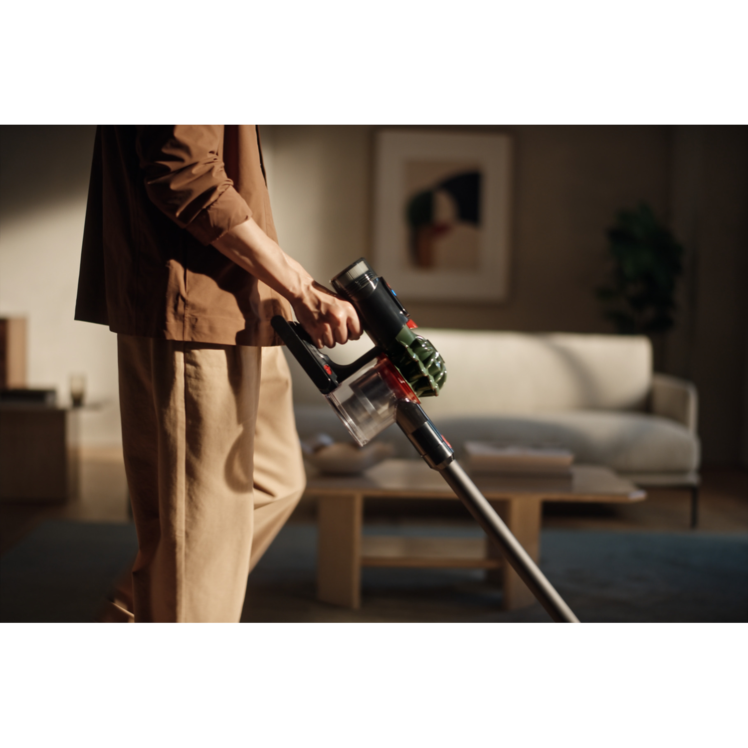 A person in brown jacket and pants vacuums a room. A modern vacuum cleaner is held, with a sofa and table in the background.