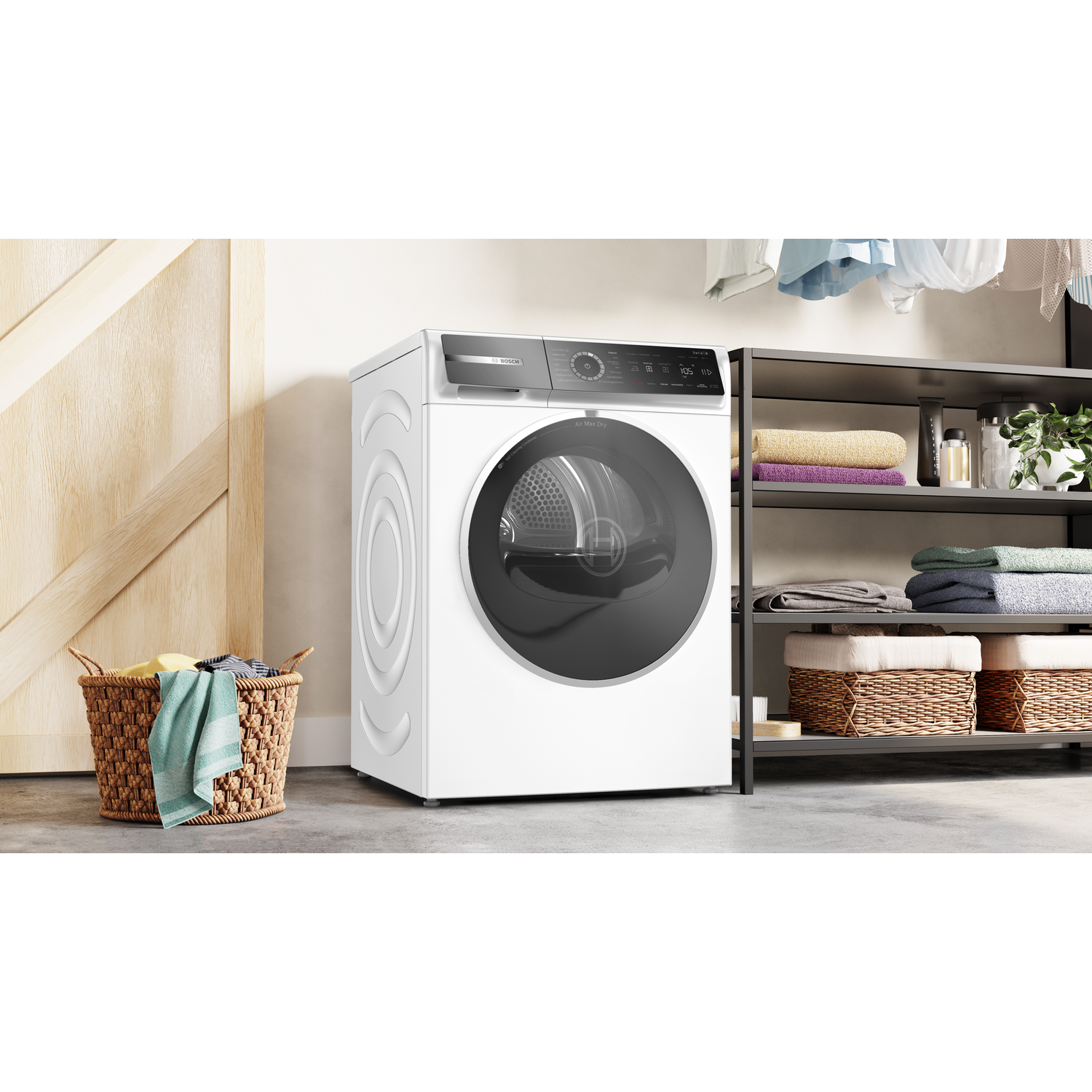 A white dryer stands next to a basket of clothes. Shelves hold folded towels. A wooden door is on the left. Clothes hang above.