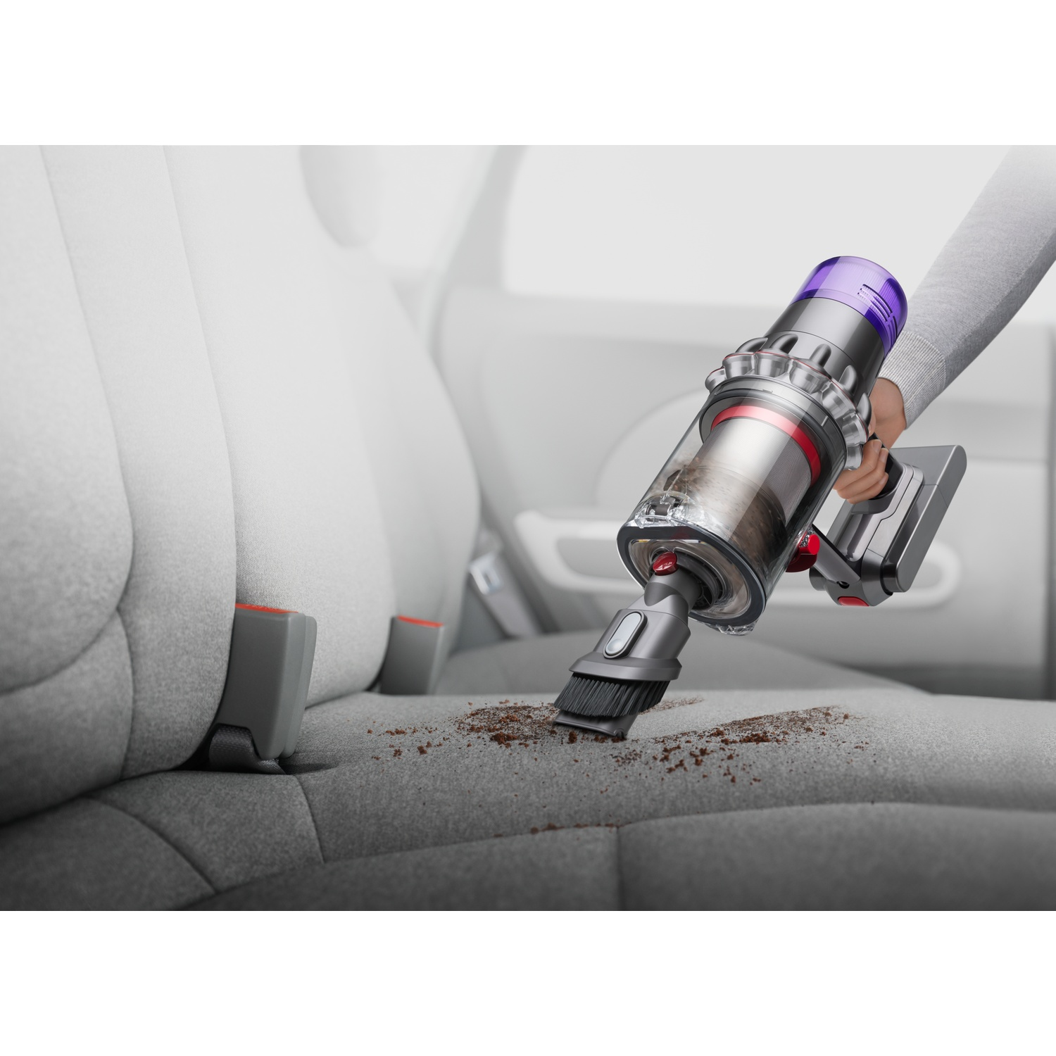 Een persoon die een autostoel stofzuigt met een Dyson. De stoel is grijs en er is vuil zichtbaar. De stofzuiger is zilver en paars.