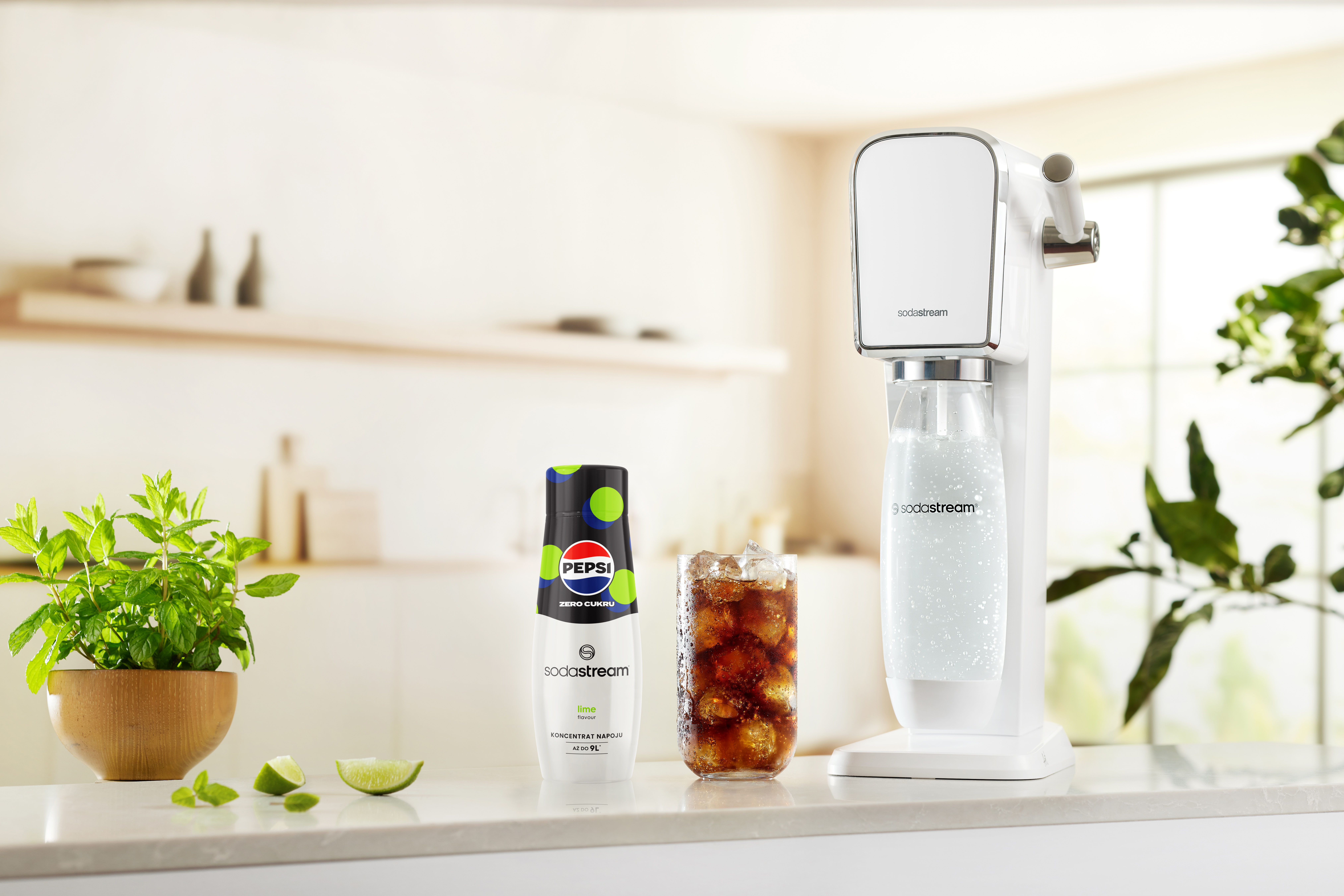 Biała maszyna SodaStream, butelka Pepsi, szklanka coli, roślina i plasterki limonki na białym blacie.