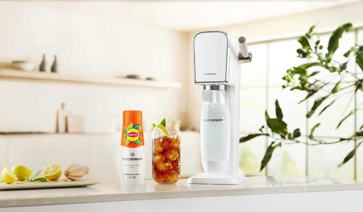 Biała maszyna SodaStream, szklanka mrożonej herbaty, butelka Lipton i cytryny na blacie z tłem kuchni.