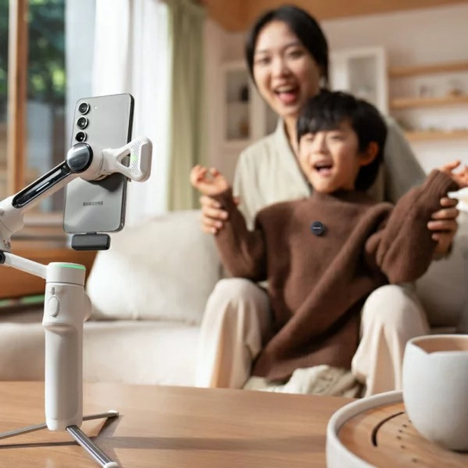 A phone on a tripod records a woman and a child smiling. The child wears a brown sweater. A white cup and tray are on the table.