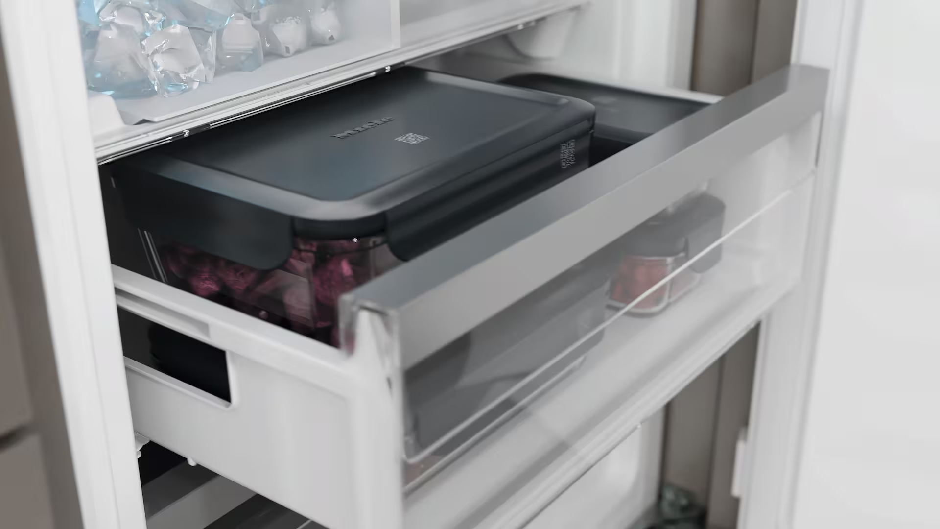 In einem Kühlschrank sind zwei Schubladen zu sehen. Die obere Schublade enthält einen schwarzen Behälter. Die untere Schublade hat Gläser mit rotem Inhalt.