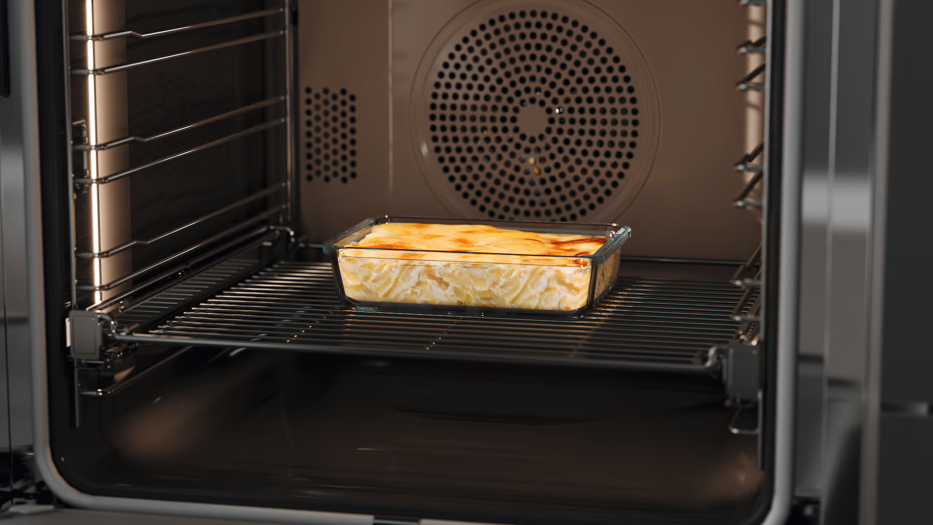 In einem Ofen befindet sich ein Glasbehälter mit gebackenem Essen auf einem Rost. Der Innenraum des Ofens ist dunkel mit einem Ventilator hinten.