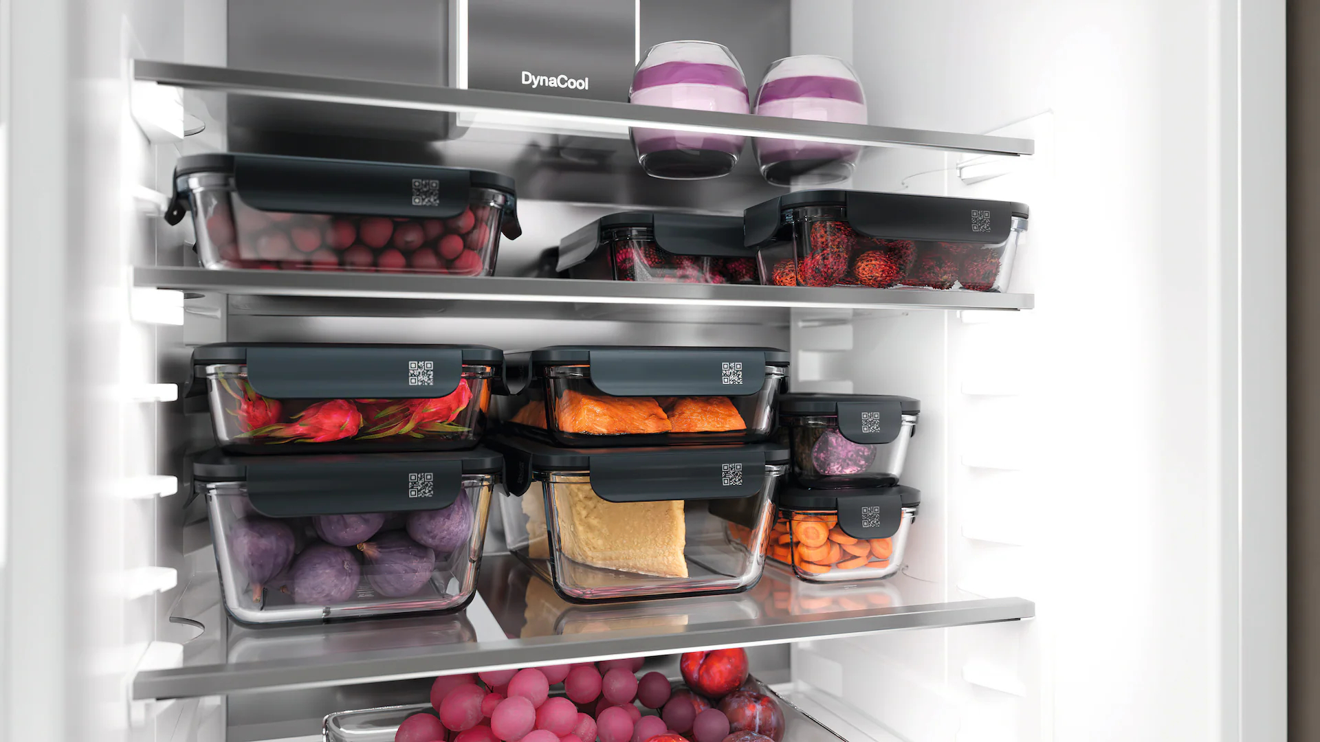 Ein Kühlschrank mit Glasbehältern voller Obst und Gemüse, darunter Trauben, Drachenfrucht und Karotten. Zwei dekorative Vasen stehen oben.