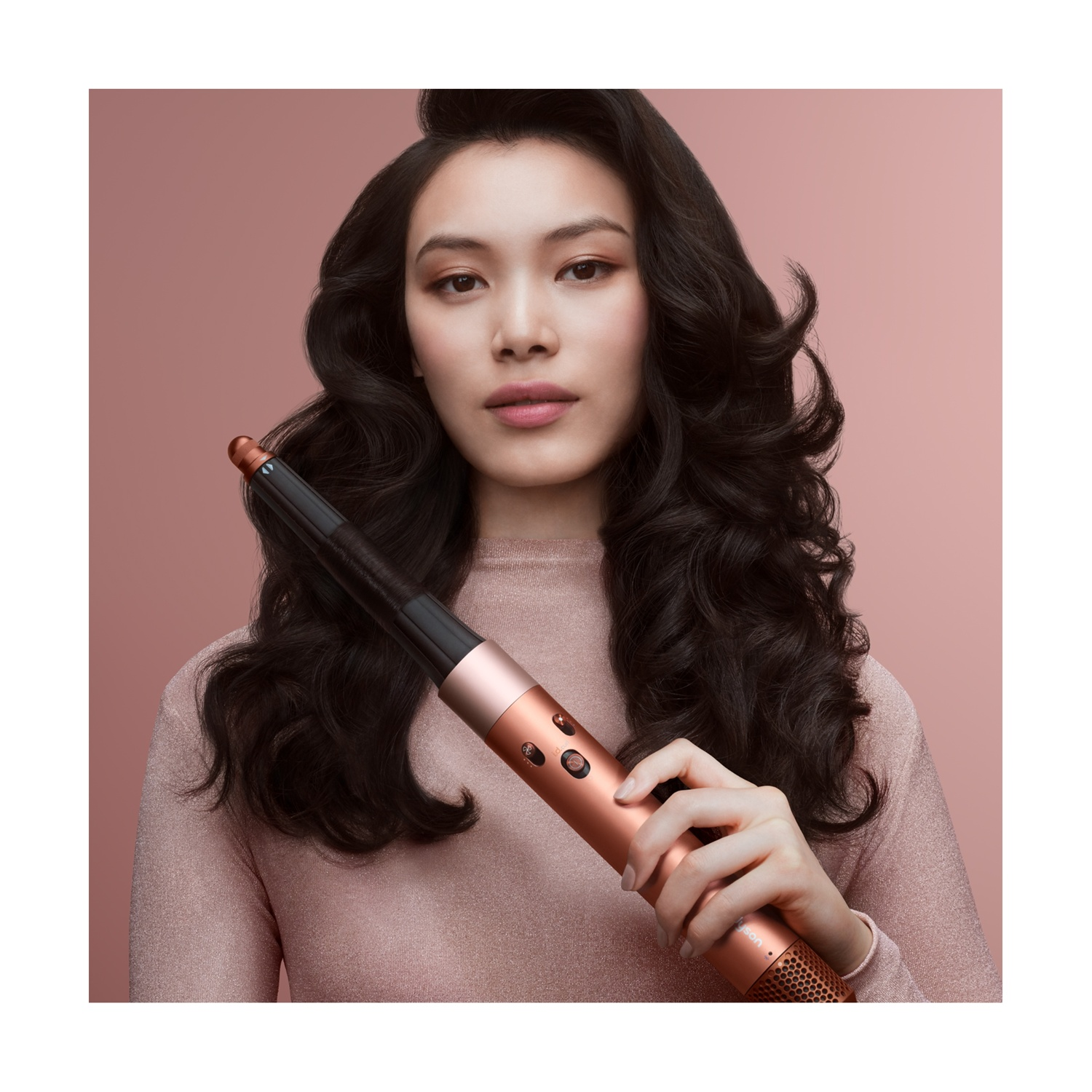 Una donna con capelli scuri e ricci tiene in mano uno strumento per lo styling dei capelli color rame. Indossa un maglione rosa su sfondo rosa pallido.