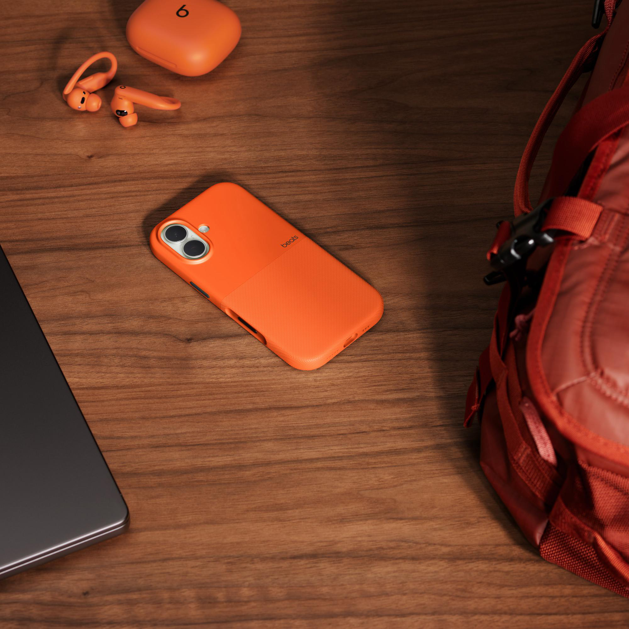 Eine orangefarbene Handyhülle, Ohrhörer und Ladecase auf Holz, mit teilweise sichtbarem roten Rucksack.