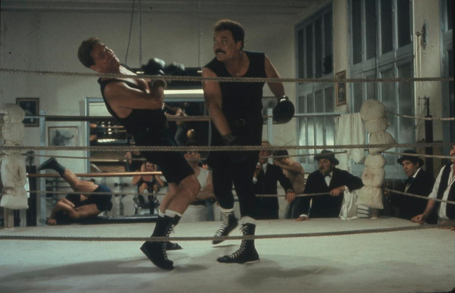 Zwei Boxer im Ring, einer am Boden. Publikum schaut zu. Boxring hat Seile und Leinwand. Männer in Anzügen im Hintergrund.