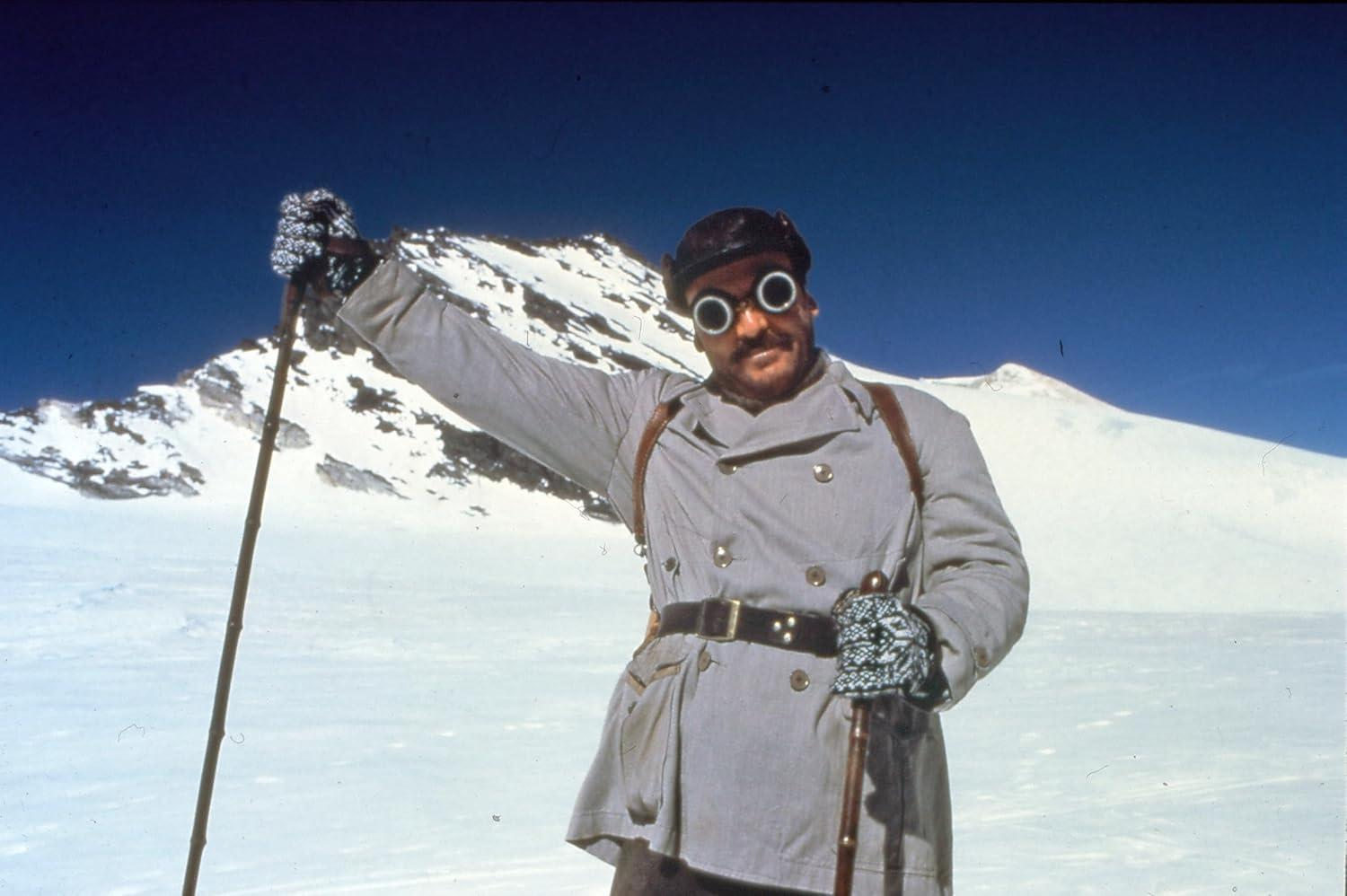 Ein Mann mit Schneegläsern und Mantel steht im Schnee und hält Skistöcke. Im Hintergrund befindet sich ein Berg unter blauem Himmel.