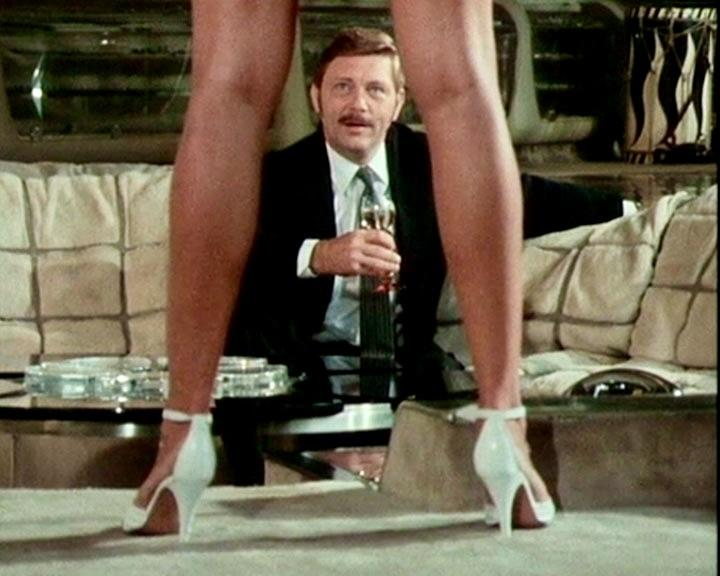 Mann im Anzug mit Glas, gesehen durch Beine einer Person in weißen Absätzen. Szene hat Couch und Tisch.