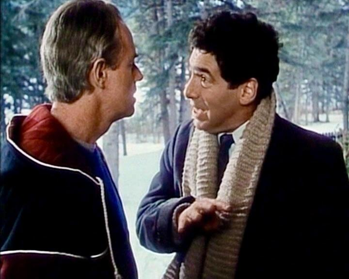 Zwei Männer unterhalten sich im Freien. Einer trägt einen Schal, der andere eine Jacke. Im Hintergrund sind Bäume zu sehen.
