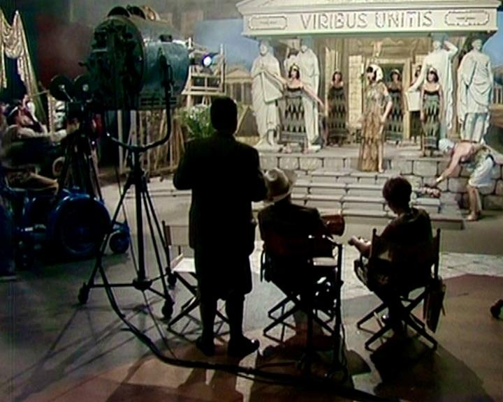 Filmset mit kostümierten Schauspielern, einer Kamera, Crewmitgliedern und einer Kulisse eines Gebäudes mit Statuen und dem Schriftzug 'VIRIBUS UNITIS'.