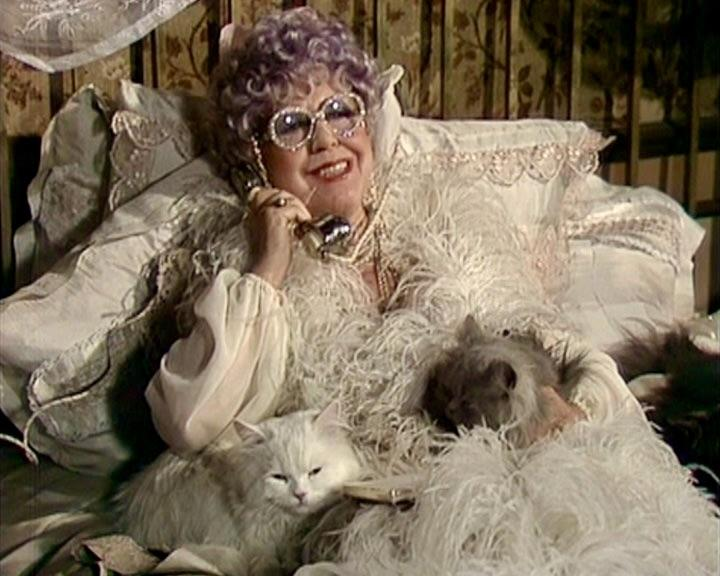 Eine ältere Frau im Bett mit Brille und Federboa, telefoniert und hat zwei Katzen.