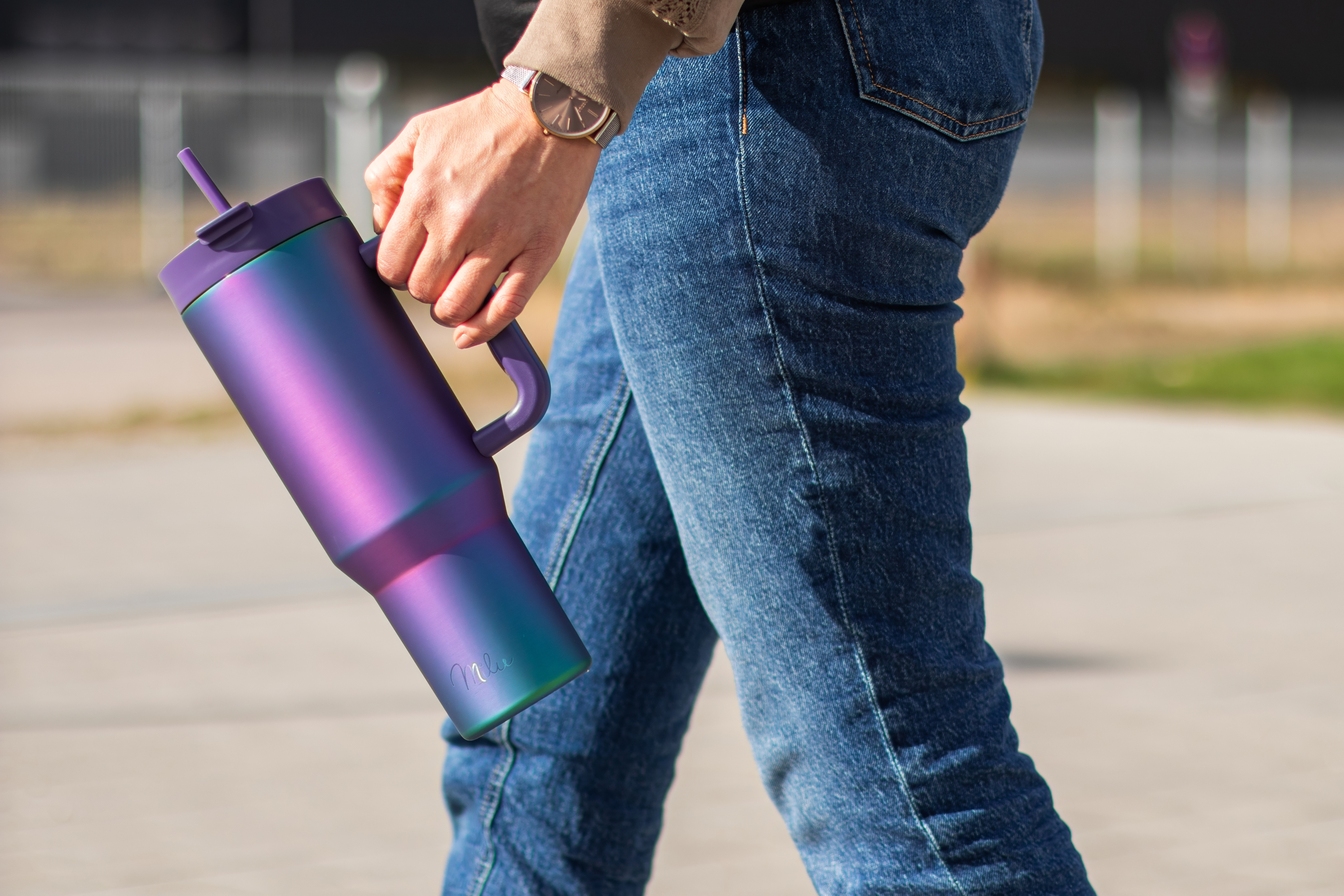 Person hält einen bunten Becher, trägt Jeans und eine Uhr. Der Hintergrund ist verschwommen mit Zaun und Grün.