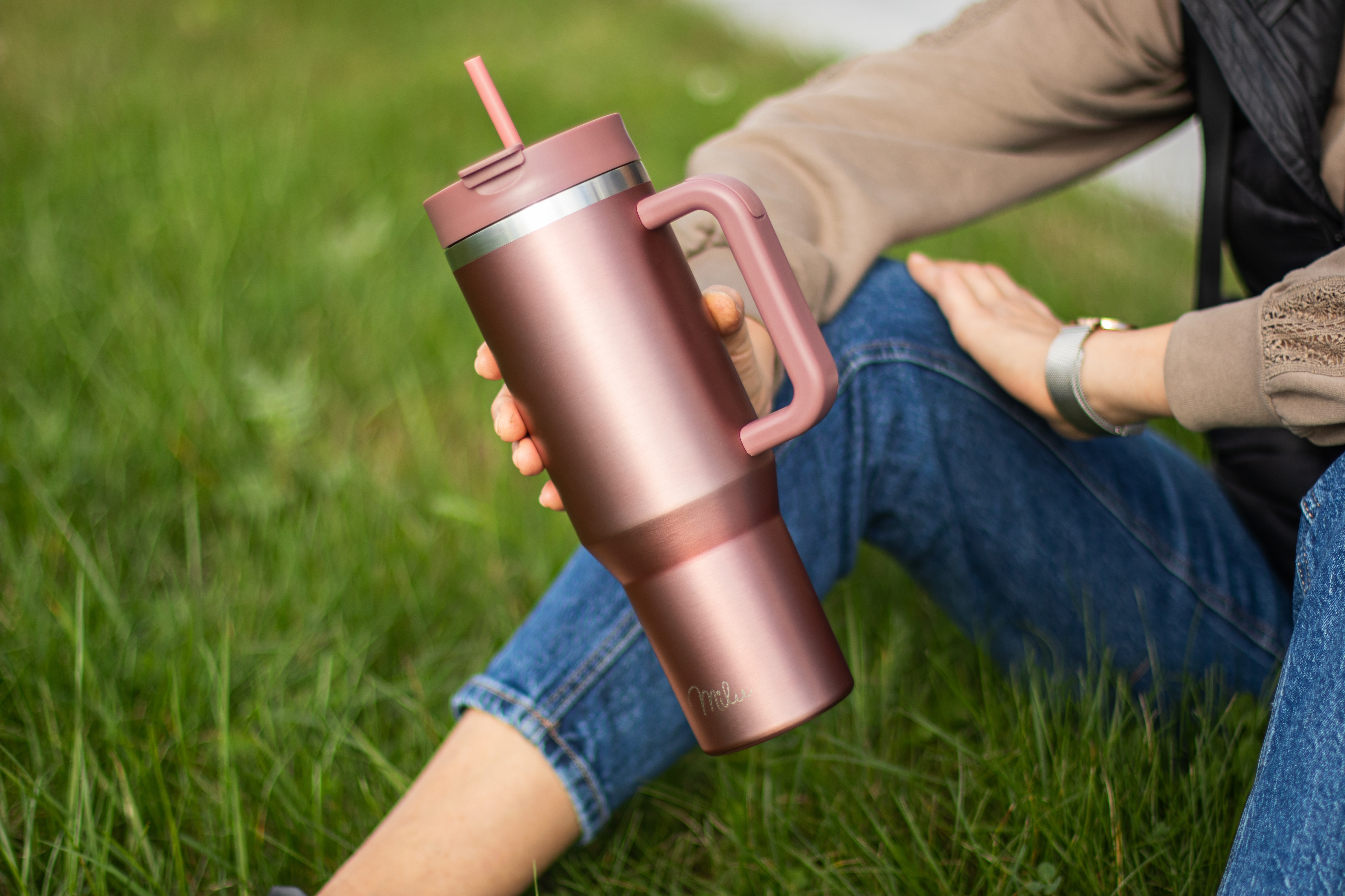 Eine Person hält einen roségoldenen Becher mit Henkel und Strohhalm, während sie auf grünem Gras sitzt.