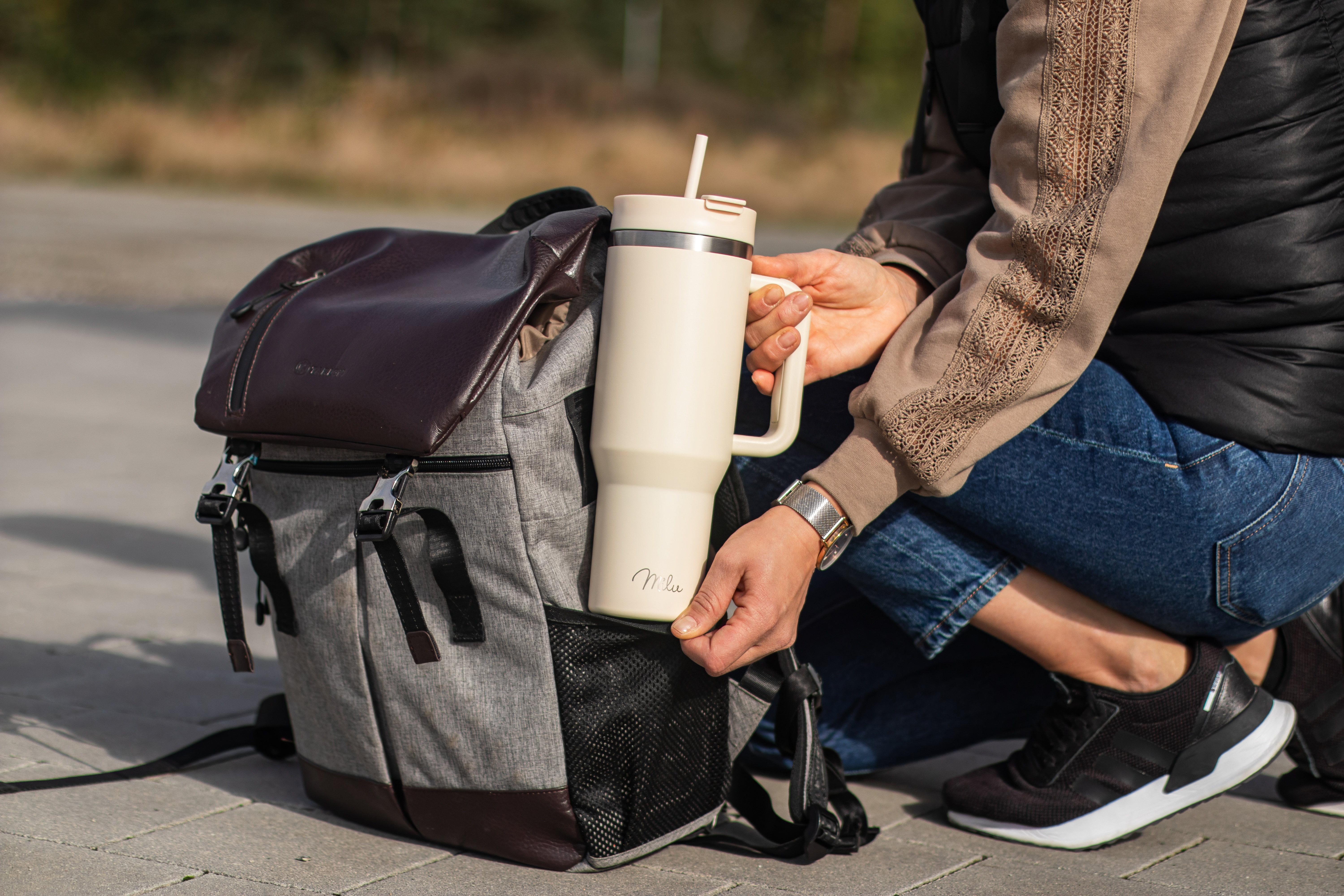 Eine Person hält einen cremefarbenen Becher neben einem grauen und braunen Rucksack. Sie trägt Jeans und ein braunes Oberteil.