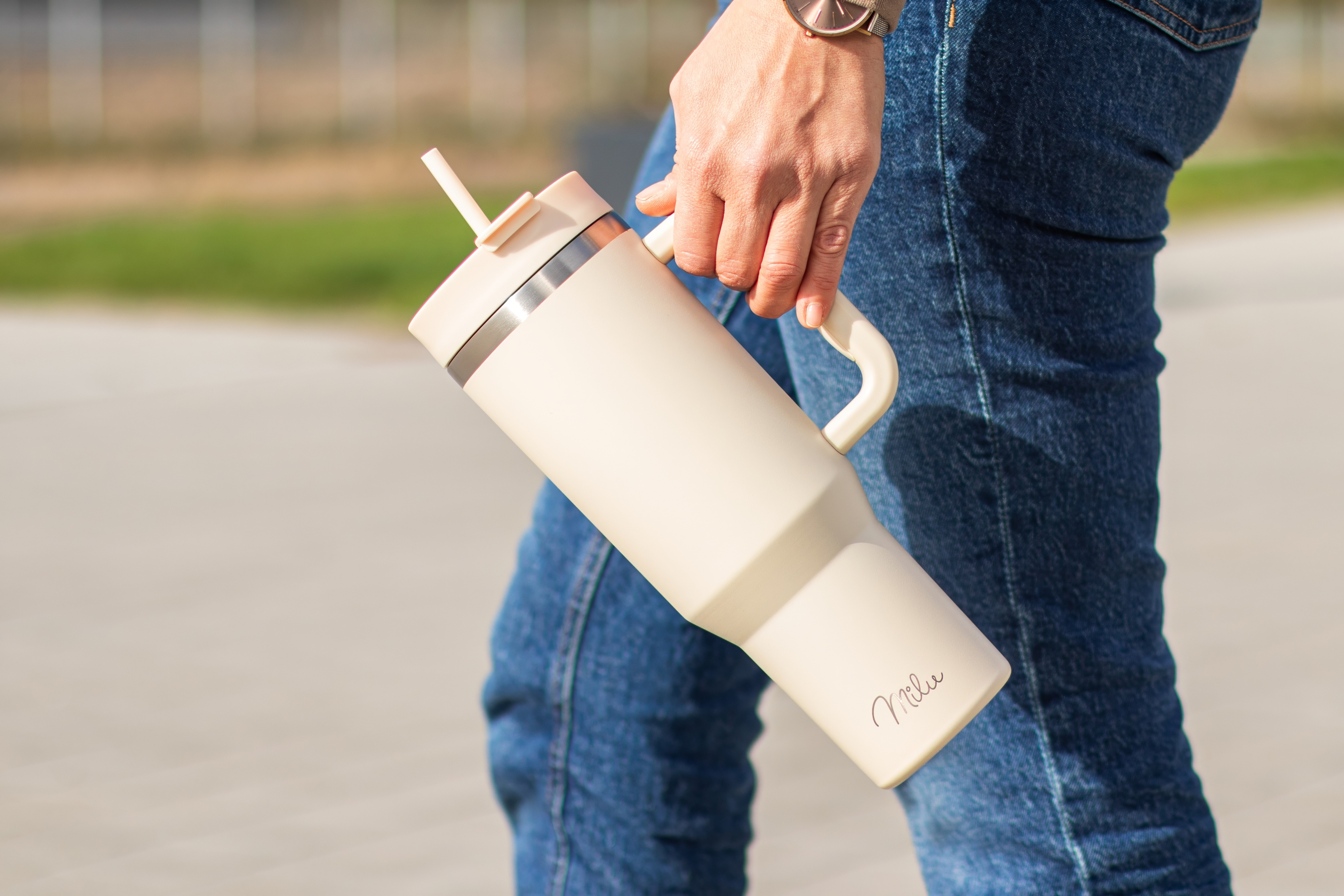 Eine Person hält einen cremefarbenen Becher mit Henkel und Strohhalm. Die Person trägt Jeans und eine Uhr.