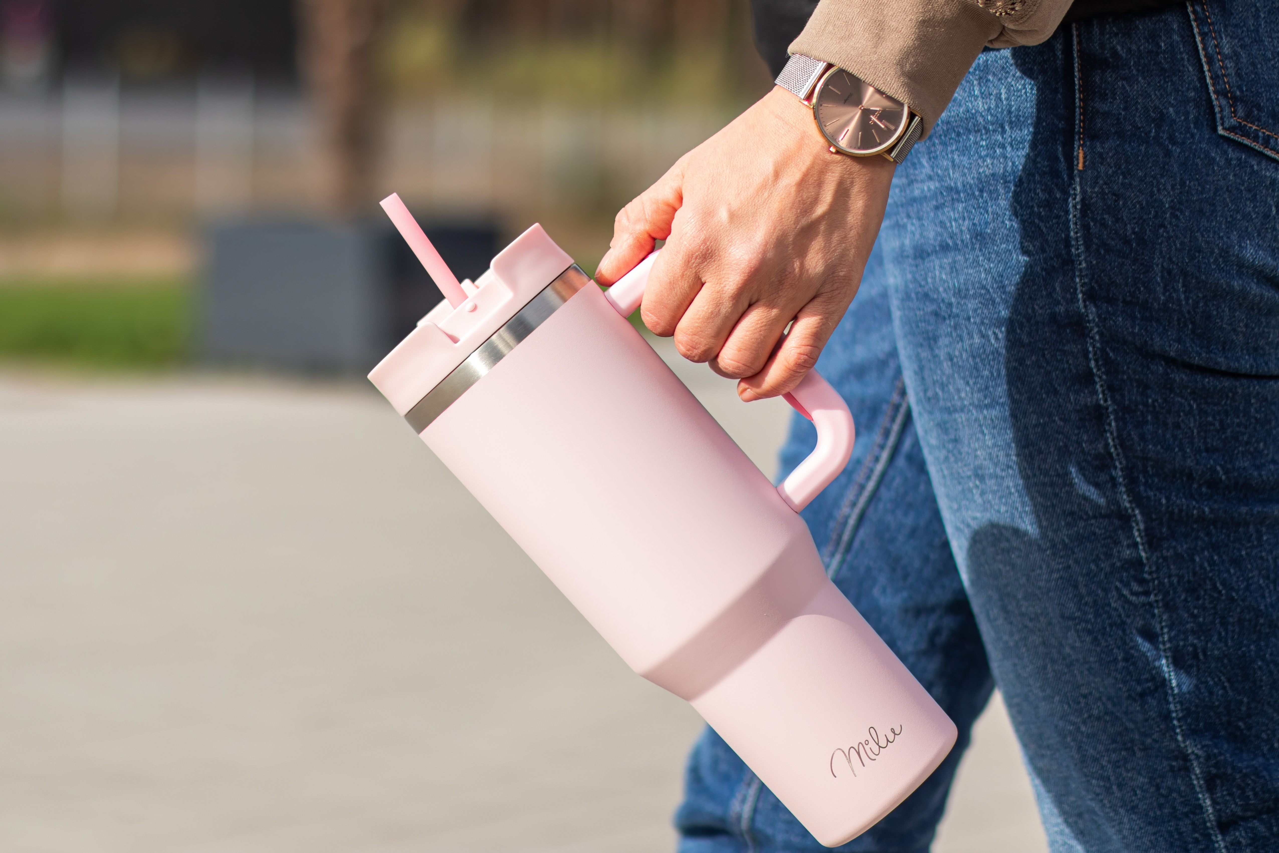 Eine Person hält einen rosa Becher mit Strohhalm. Die Person trägt eine Uhr und Jeans. Der Hintergrund ist unscharf.
