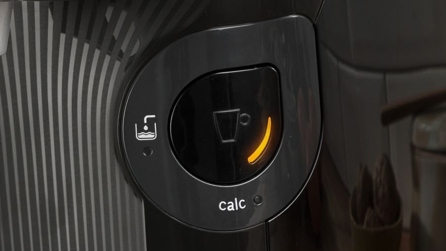 Primer plano del botón negro de una cafetera. Tiene un icono de taza y la palabra 'calc'. Una luz naranja ilumina el botón.