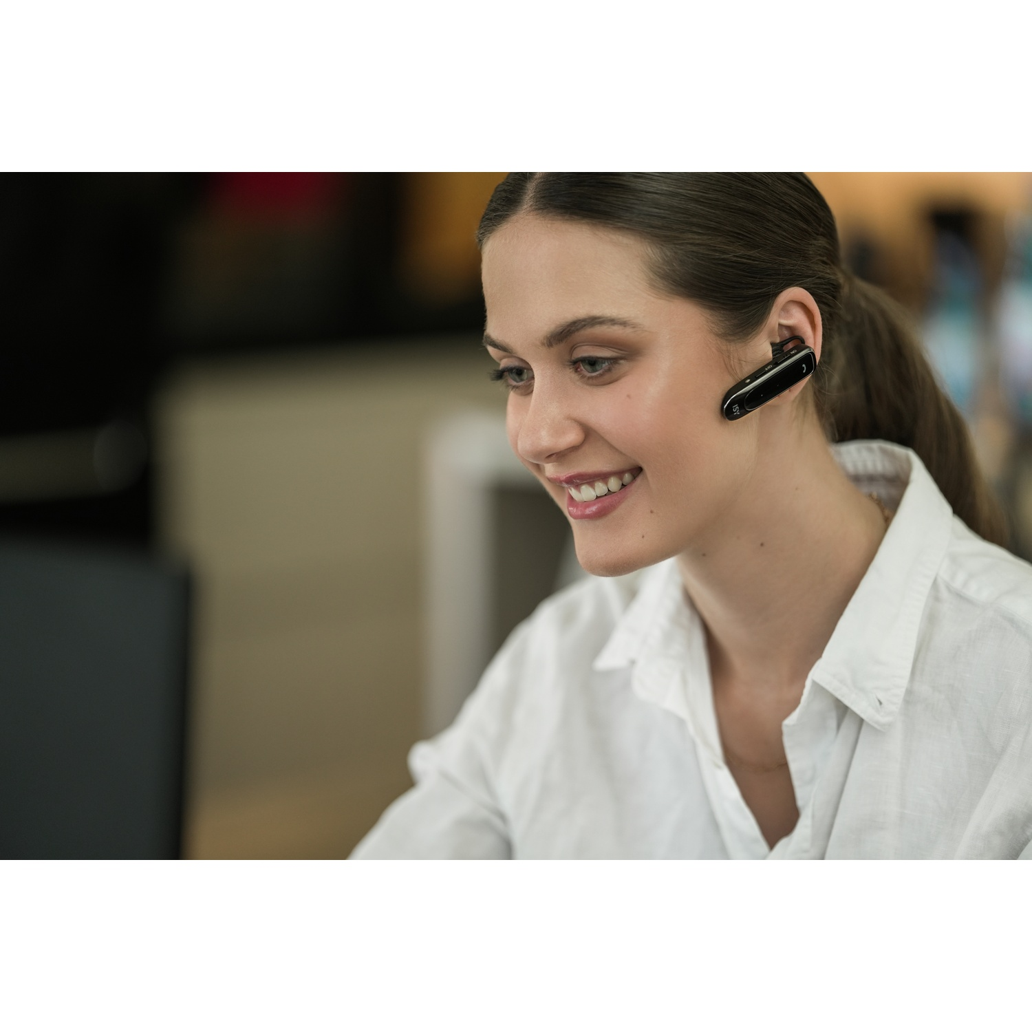 A smiling woman wearing a white shirt and a headset, looking at a screen. The background is blurred.