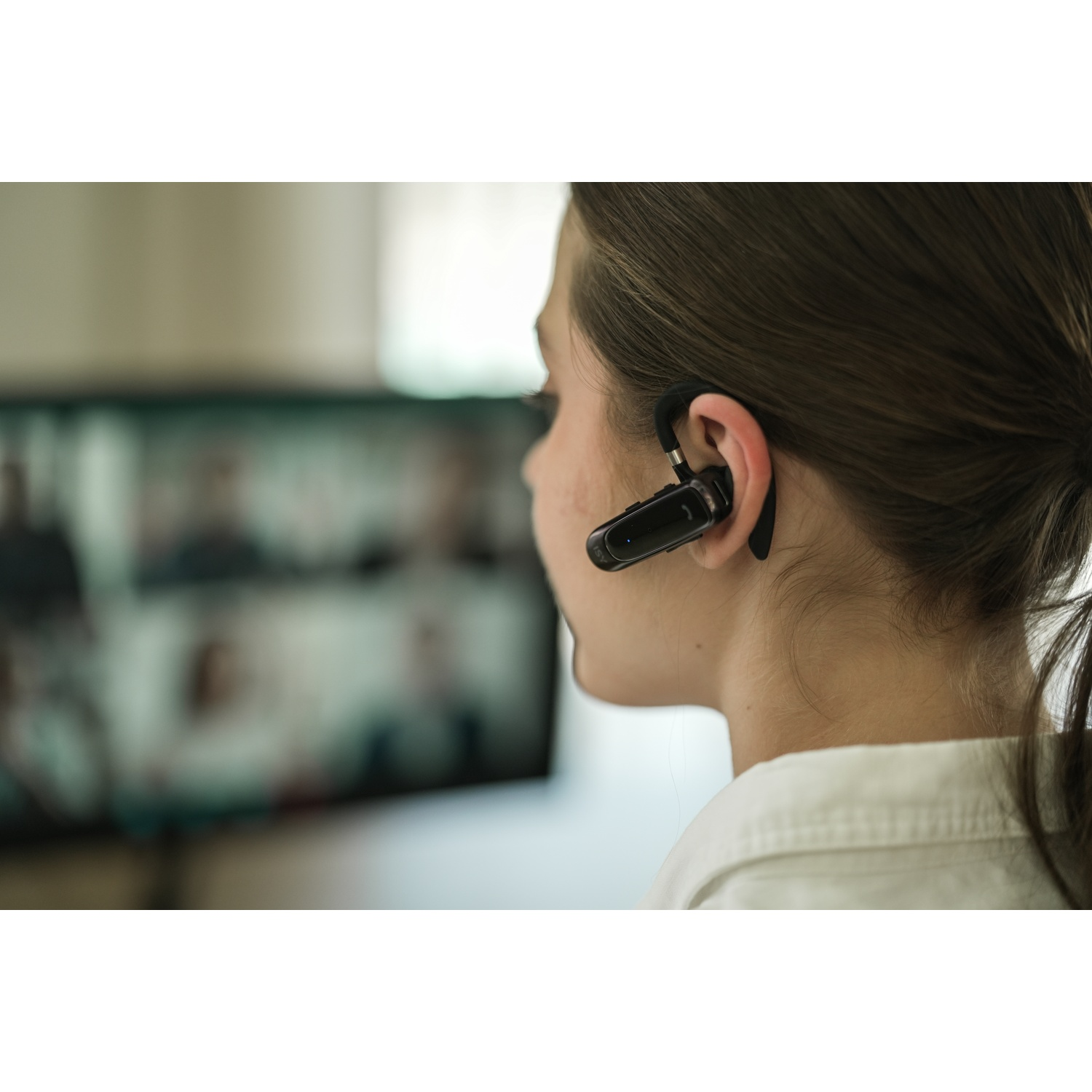 A woman wearing a headset. She has dark hair and is looking at a screen with people on it. She is wearing a white shirt.