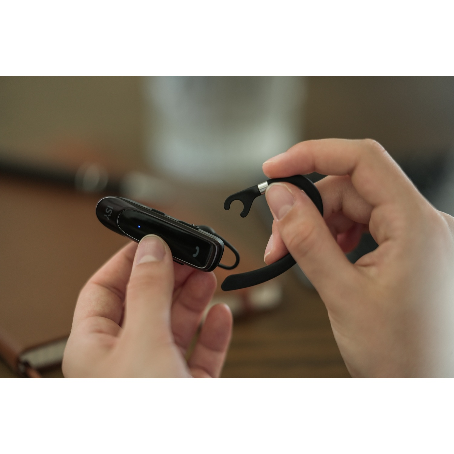 Hands holding a black Bluetooth headset. The headset is being assembled. Background has a brown table and blurred wall.