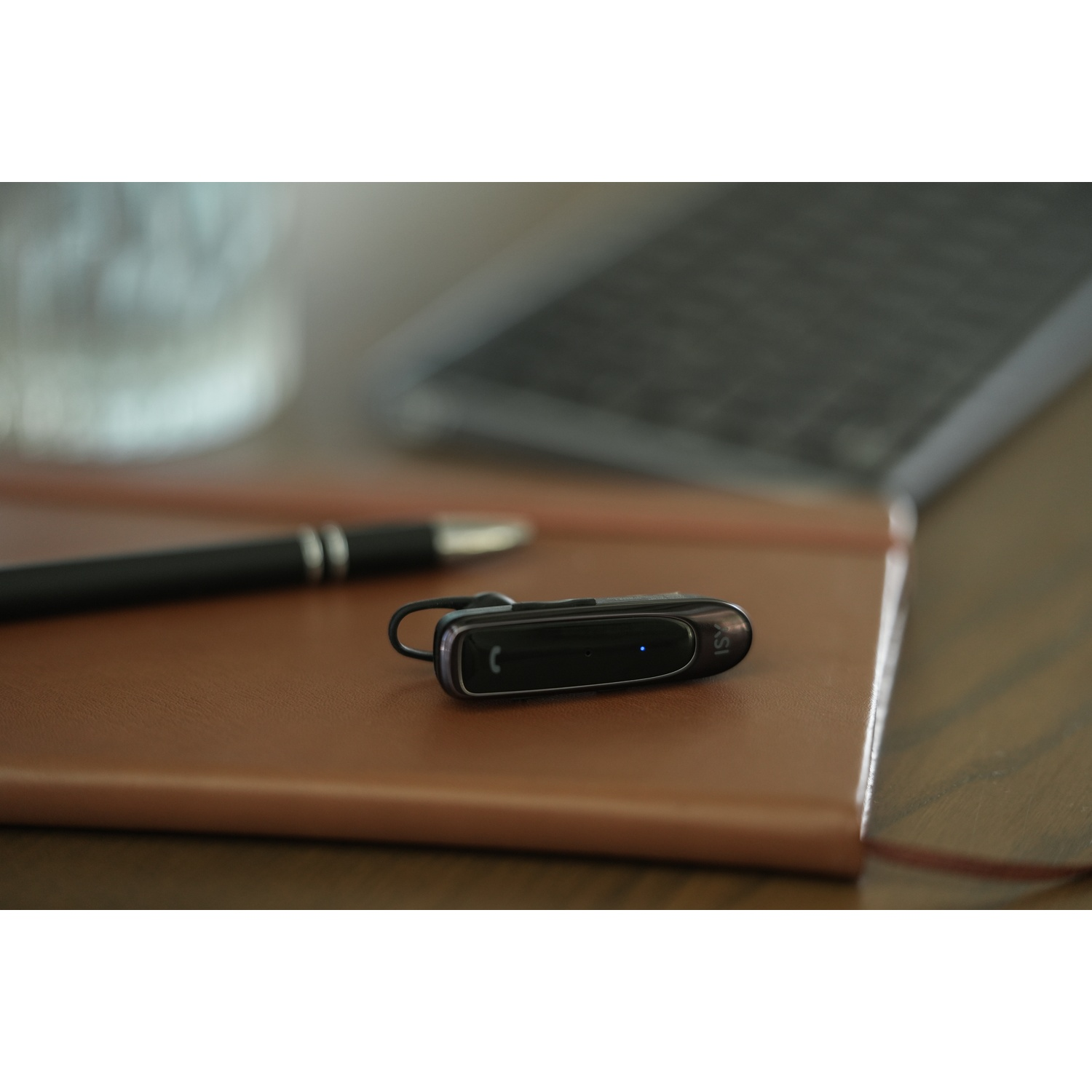 A black Bluetooth headset lies on a brown leather notebook. A pen and a tablet are also visible. The background is blurred.