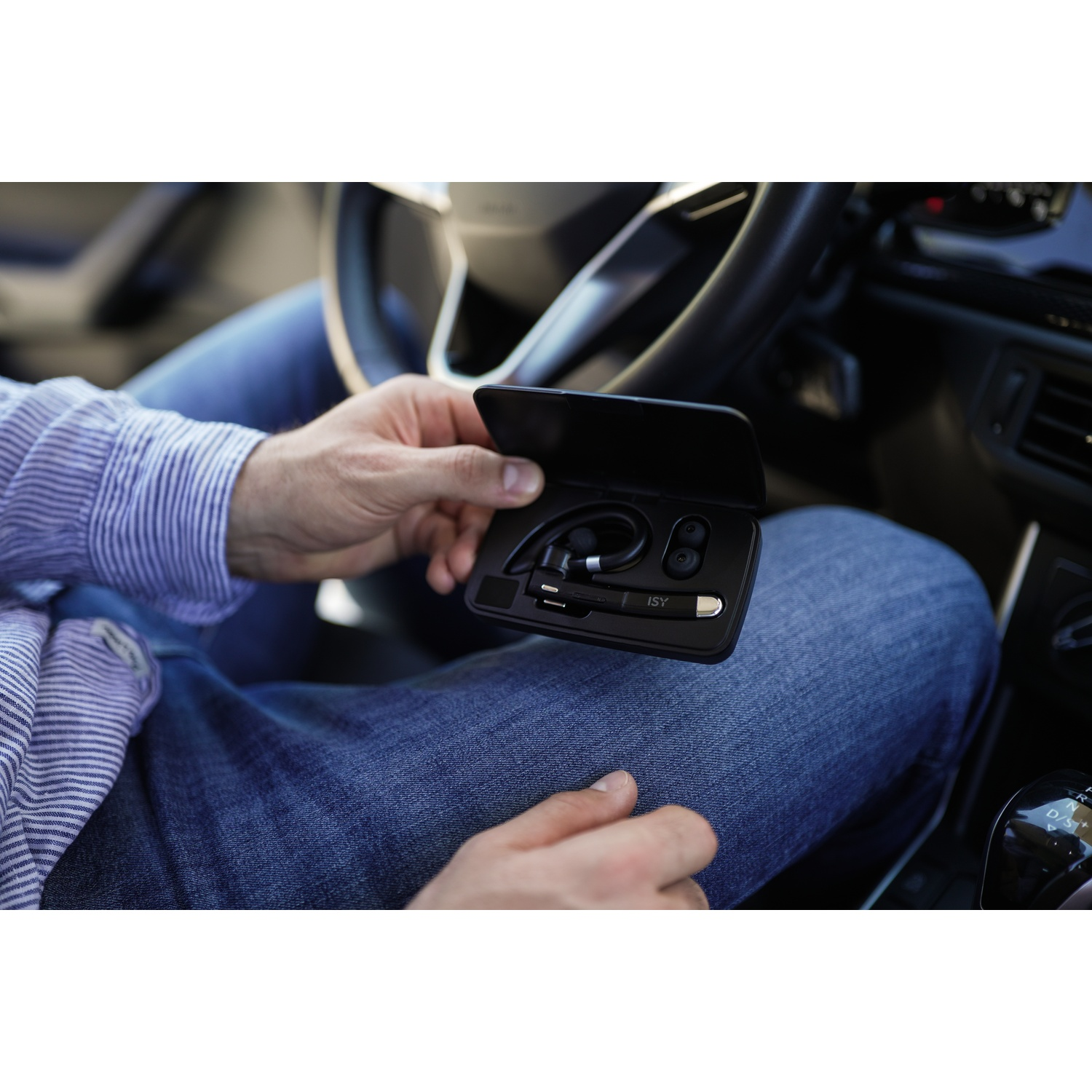 A person in a car holds a black headset case. The person wears a blue striped shirt and jeans. Steering wheel is in the background.
