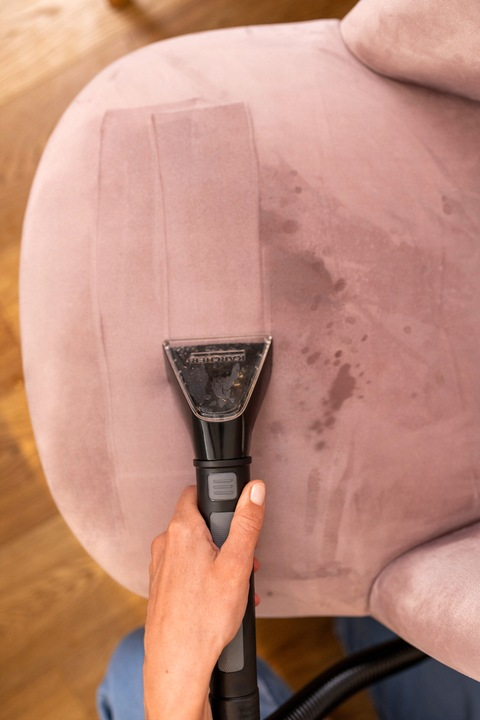 Ręka odkurza różowe krzesło. Głowica odkurzacza jest czarna. Krzesło jest poplamione, widoczna podłoga.