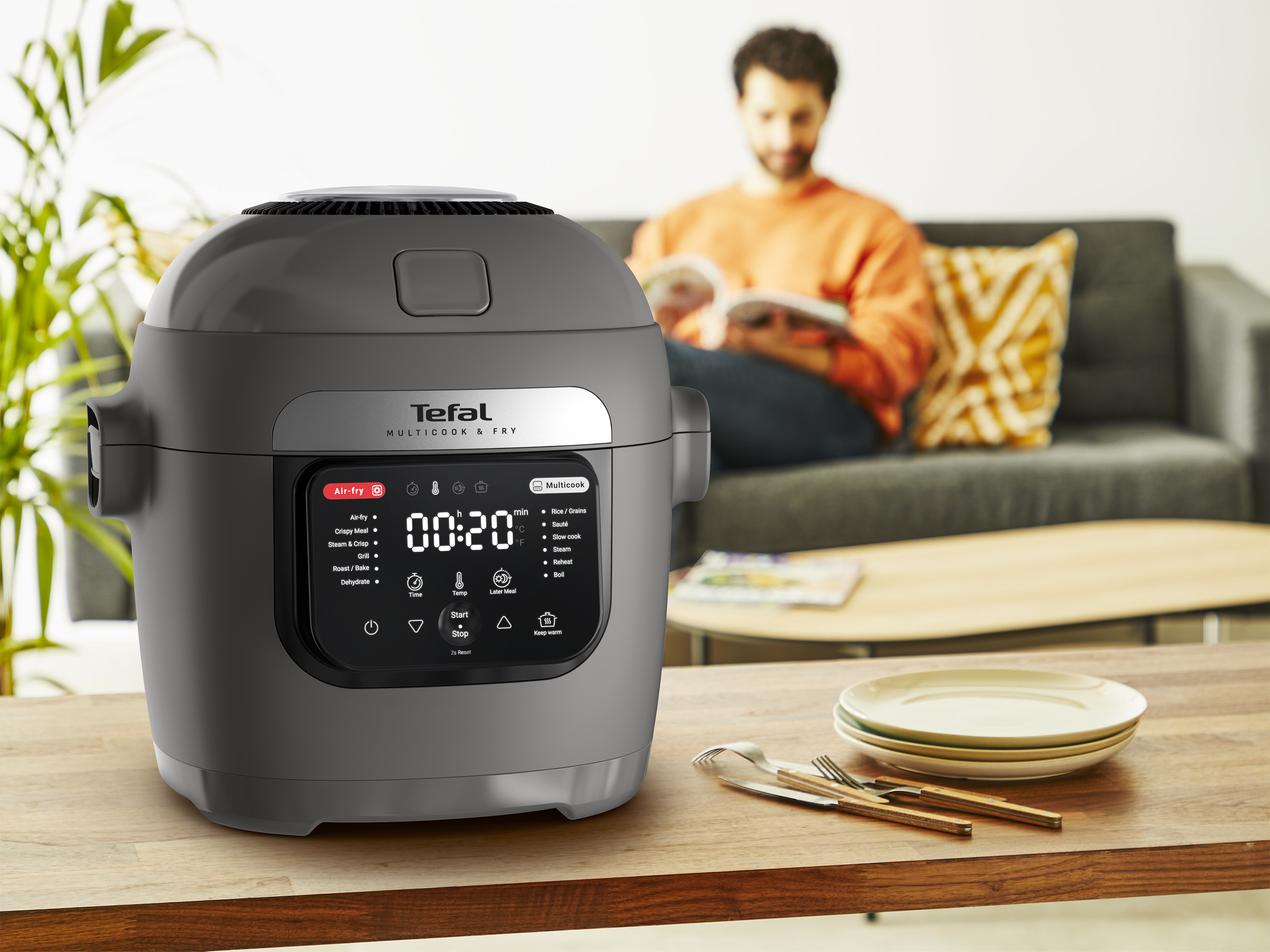 Tefal multicooker faasztalon. Egy személy olvas a kanapén a háttérben. Tányérok és evőeszközök vannak.