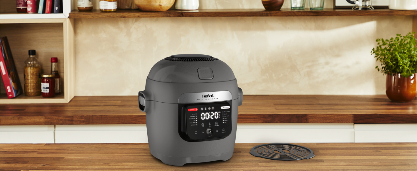 Egy szürke Tefal multifőző egy fa pulton áll. Egy fém grill és egy növény is jelen van. A háttérben polcok üvegekkel.
