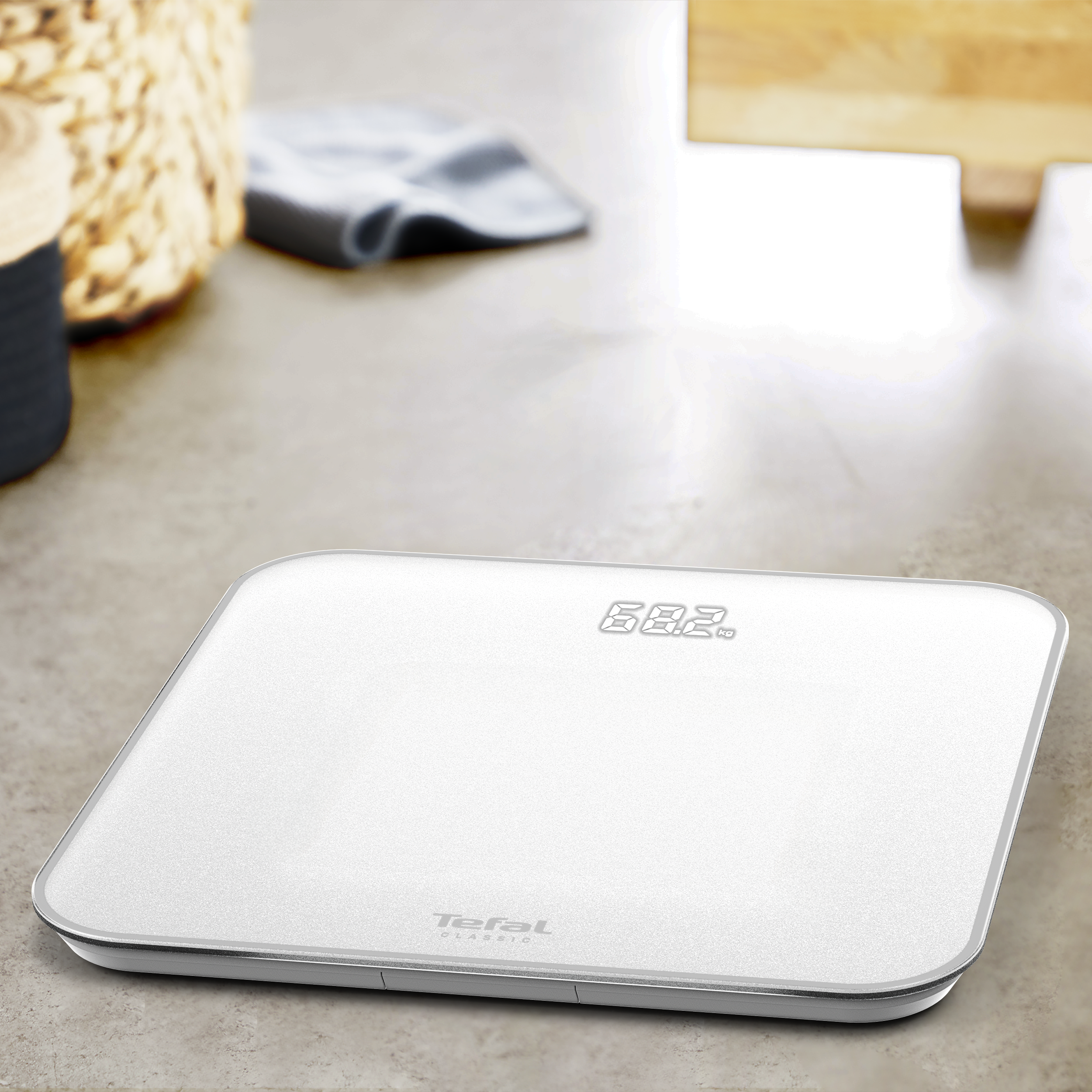 Egy fehér Tefal Classic digitális mérleg 68,2 kg-ot mutat. Kosár, szürke törölköző és fa a háttérben.