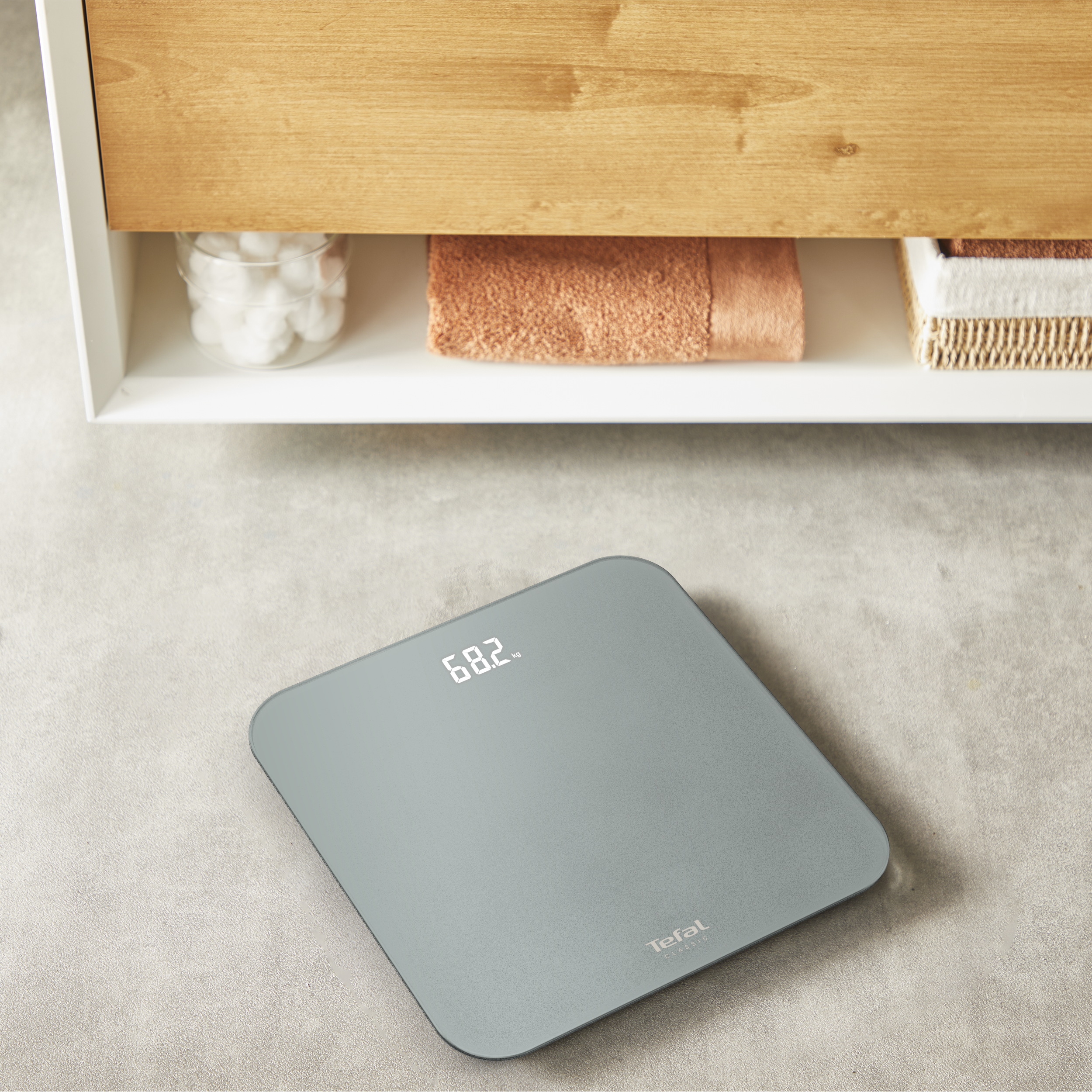 Egy digitális mérleg 68,2 kg-ot mutat. A felette lévő polcon törölköző, vattagolyók és kosarak vannak.
