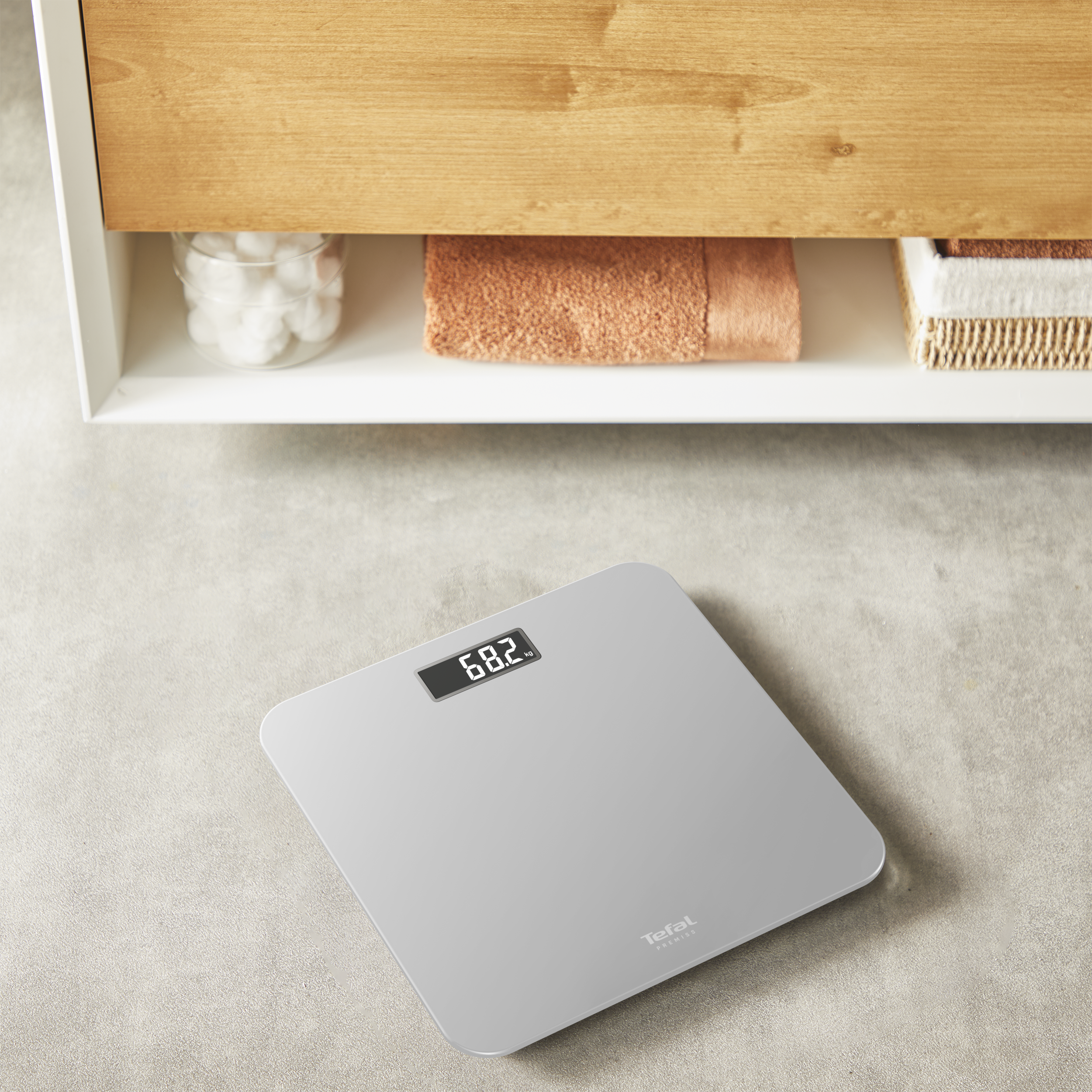 Egy digitális mérleg 68,2 kg-ot mutat. Felül egy fa szekrény, törölközővel és vattagolyókkal látható.