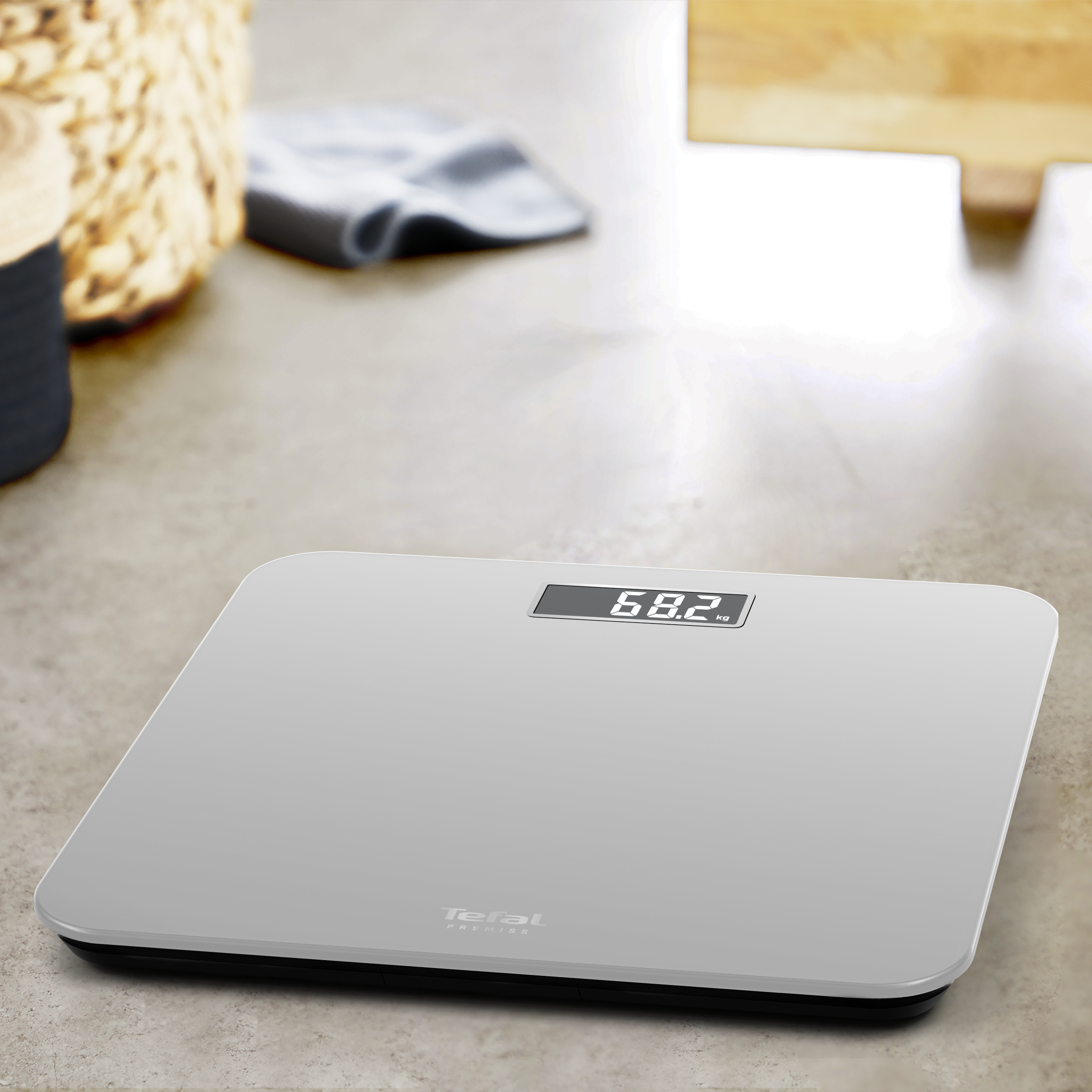 Egy digitális mérleg 68,2 kg-ot mutat. Egy szürke mérleg egy pettyes felületen nyugszik. Egy kosár és egy törölköző elmosódott a háttérben.