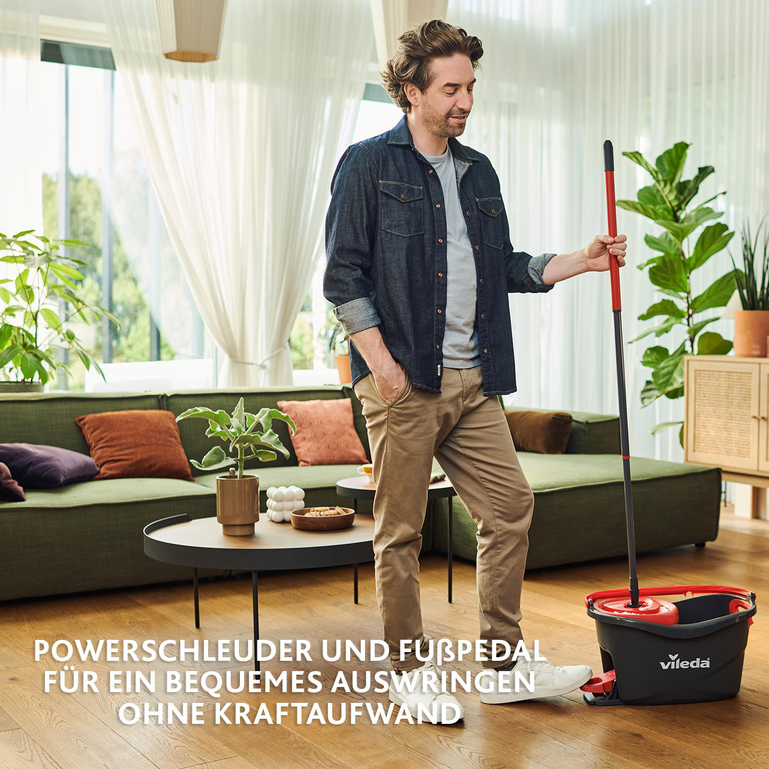 Mann mit Wischmopp und Eimer. Grüne Couch und Pflanzen im Hintergrund. Text auf dem Boden: 'Powerschleuder und Fußpedal'.