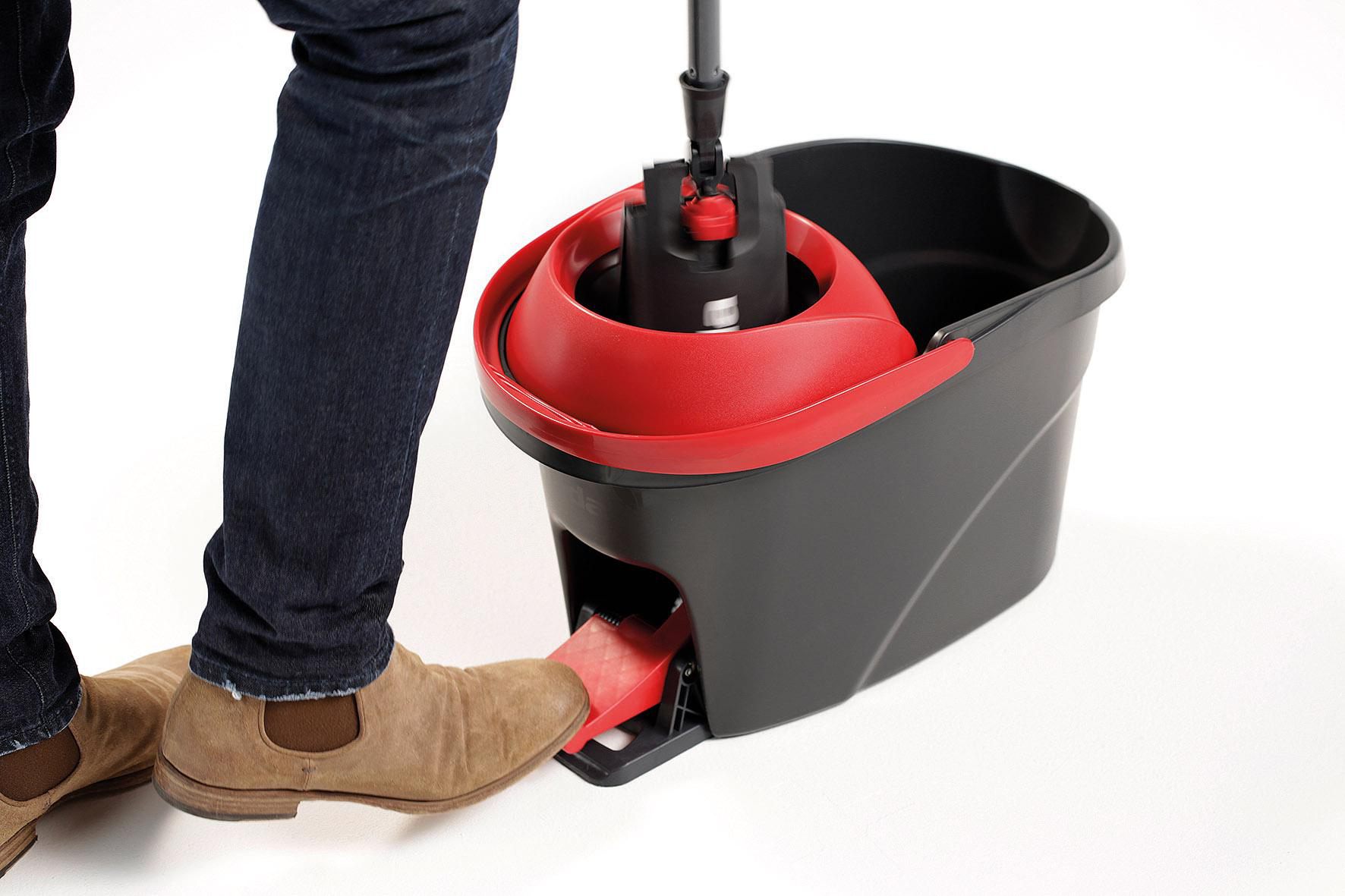 Eine Person tritt auf ein rotes Pedal eines schwarz-roten Wischeimers. Die Person trägt Jeans und braune Schuhe.