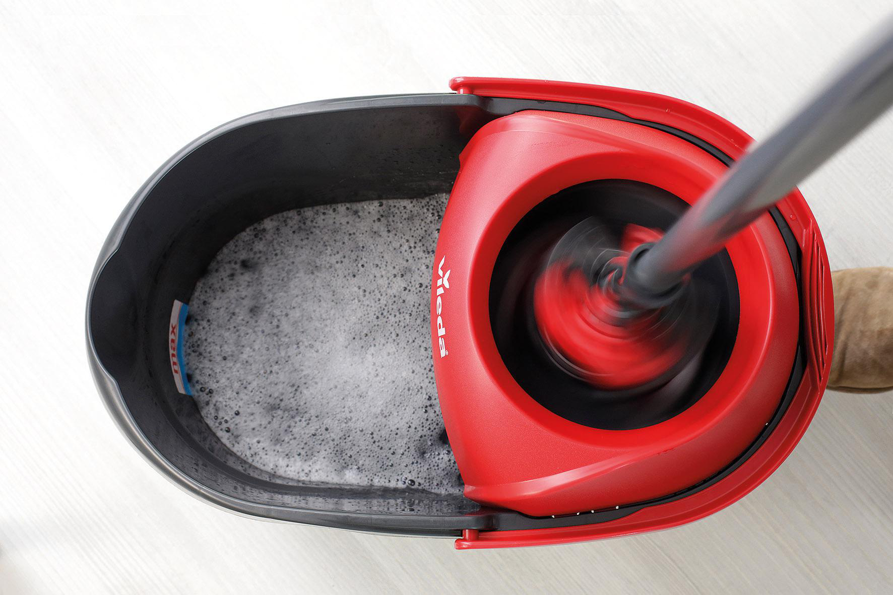 Ein roter und schwarzer Vileda-Wischer wird in einem schwarzen Eimer mit Seifenwasser verwendet. Der Wischer ist in Bewegung.