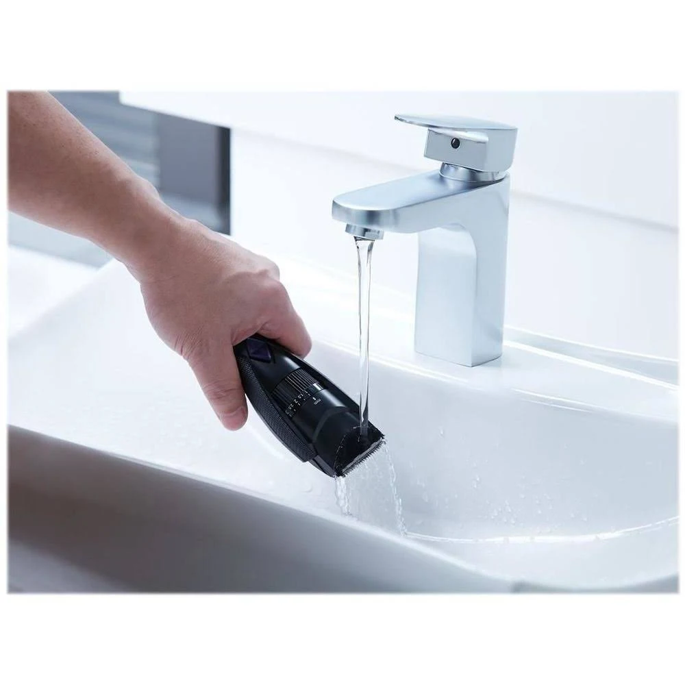Una persona lava una cortadora de cabello negra bajo agua corriente en un lavabo blanco. Un grifo plateado está arriba.