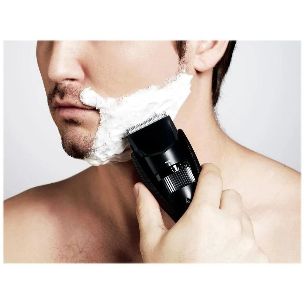 Un hombre se afeita la barba con una afeitadora eléctrica. Crema de afeitar blanca cubre la mitad de su cara. La afeitadora es negra.