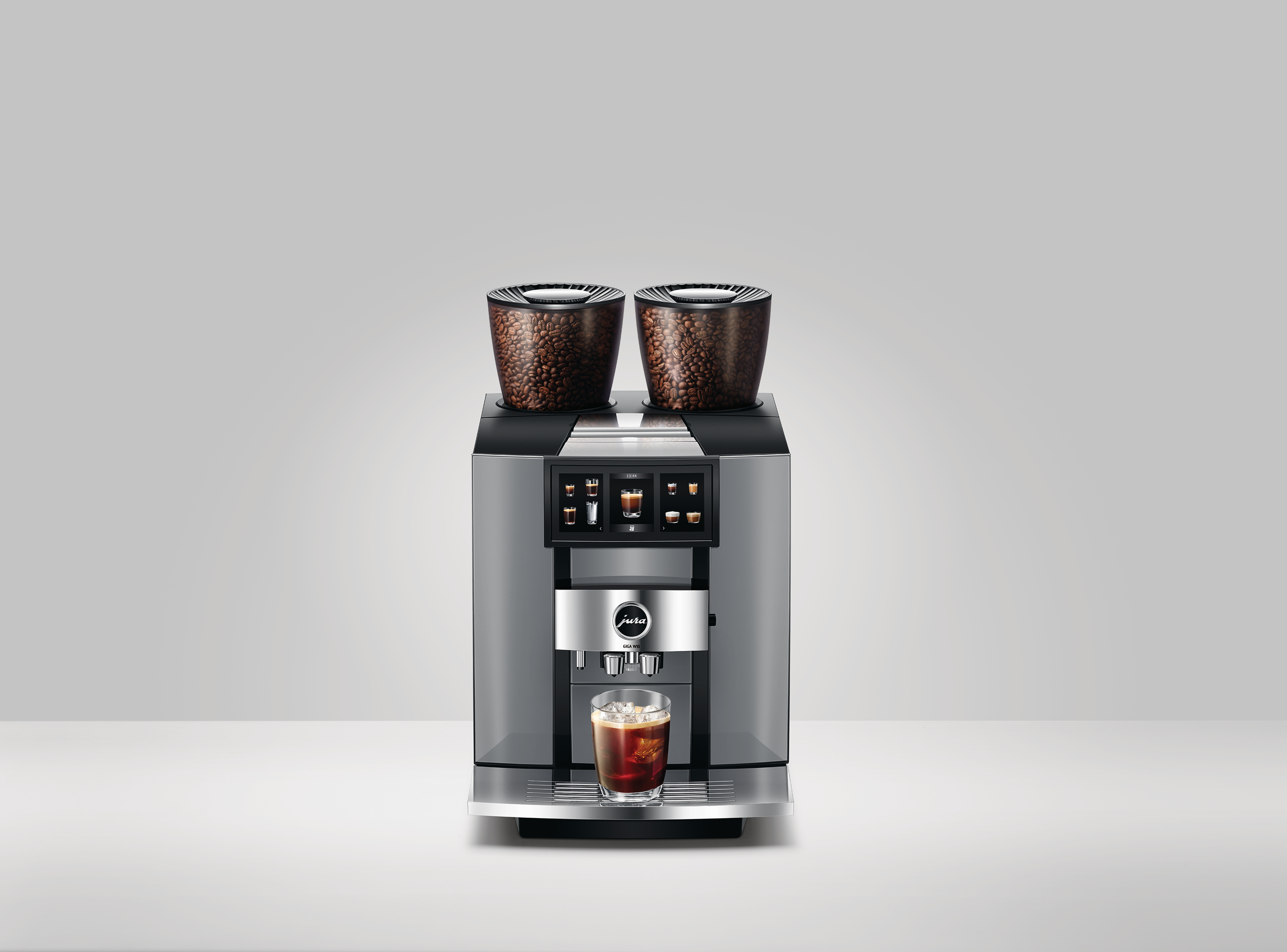 Eine moderne Kaffeemaschine mit zwei Bohnenbehältern. Ein Glas Kaffee mit Eis steht unter der Maschine. Der Hintergrund ist weiß.