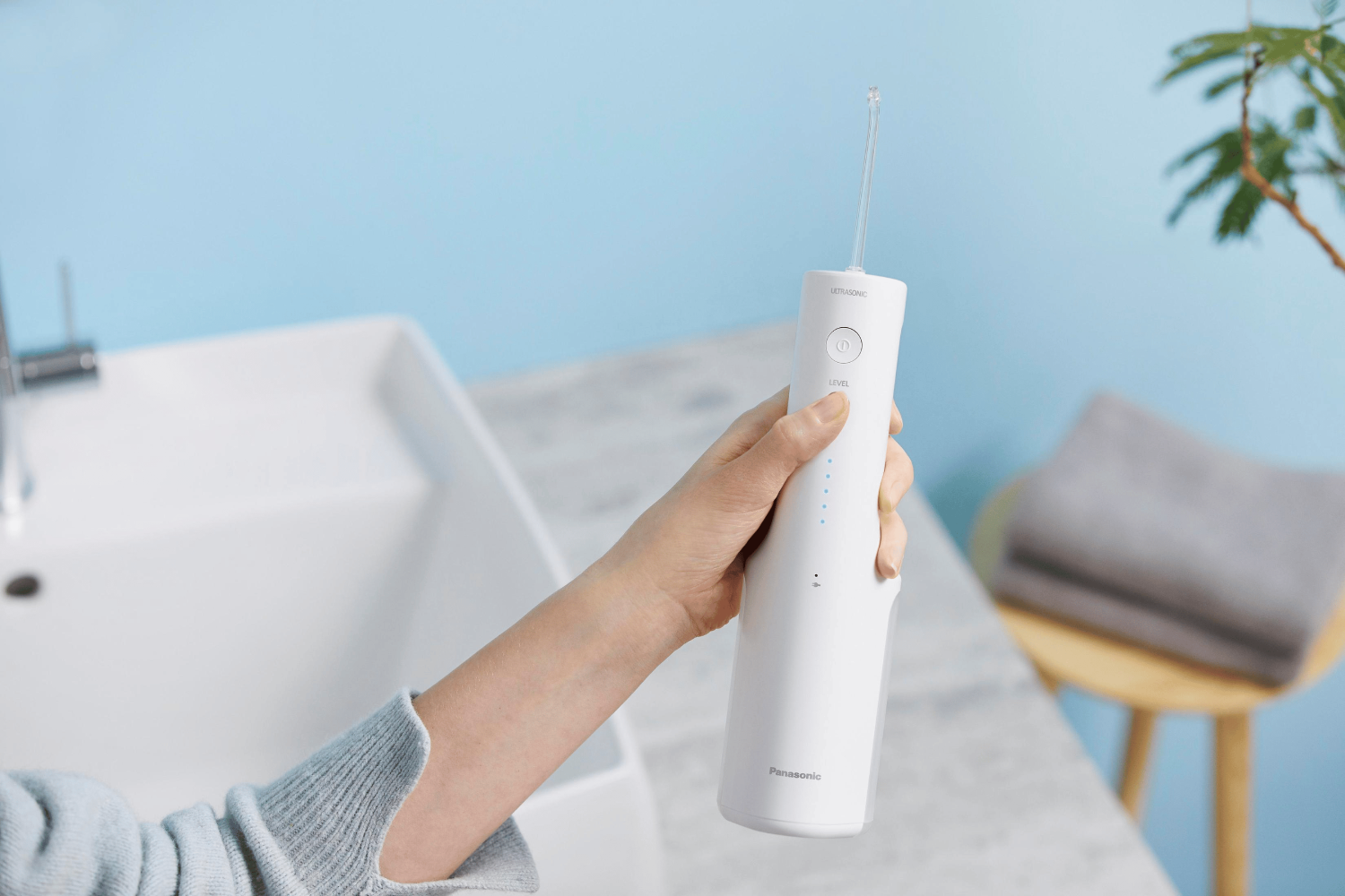 Una mano sostiene un irrigador bucal Panasonic blanco con luz azul. Un lavabo y una pared azul al fondo.