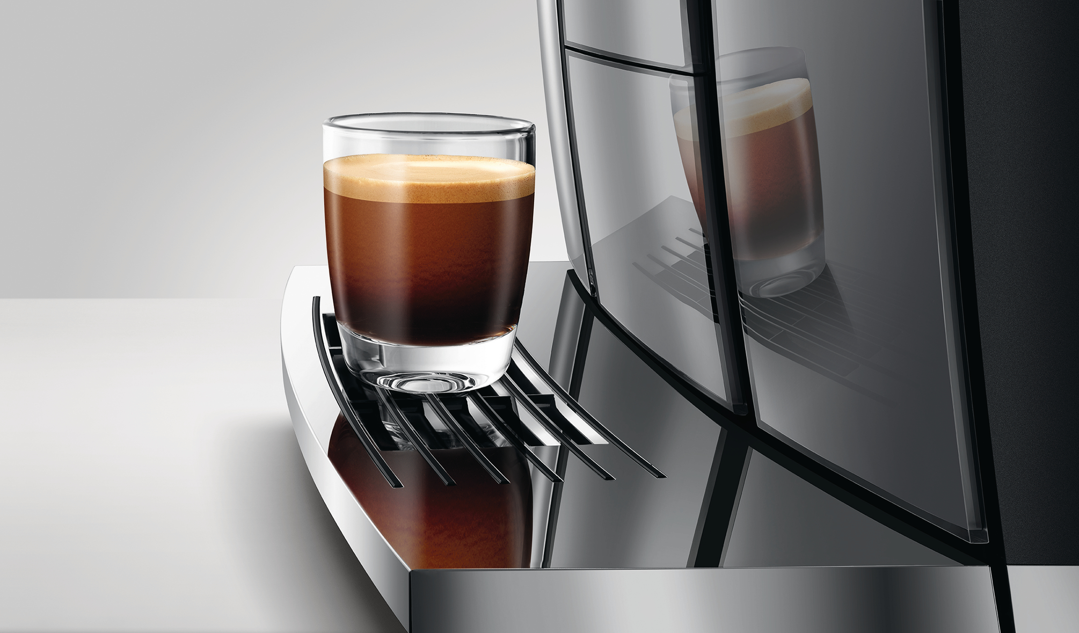 Ein Glas Espresso mit Crema steht auf einer Kaffeemaschine. Die Maschine hat ein schwarz-silbernes Design, das Glas reflektiert.