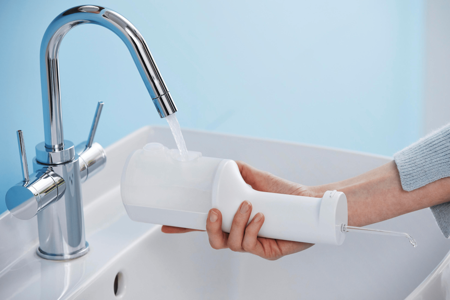 Una persona sostiene un irrigador oral blanco debajo de un grifo con agua fluyendo, encima de un lavabo blanco.