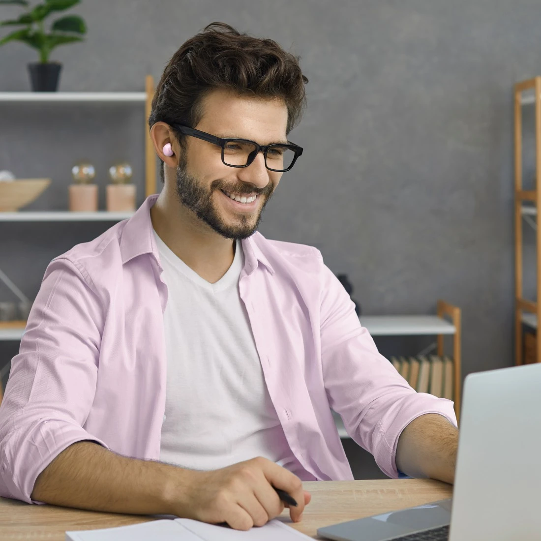 Egy mosolygó férfi szemüveggel és fülhallgatóval laptopon dolgozik. Rózsa inget visel. Egy jegyzetfüzet és egy toll van az asztalon.