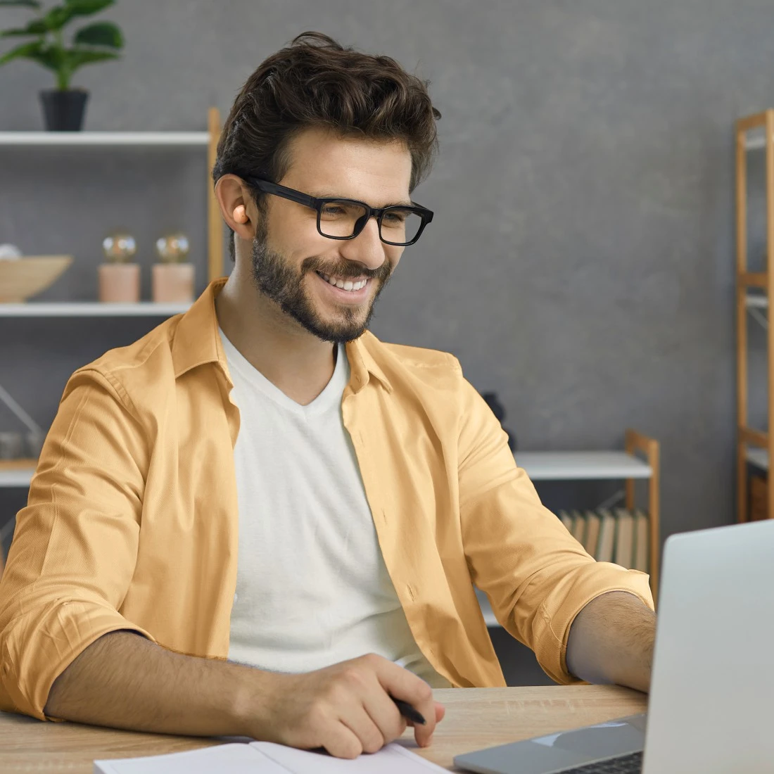 Egy mosolygó férfi szemüveggel laptopon dolgozik. Sárga inget és fehér pólót visel. A desk-en egy jegyzetfüzet és egy toll található.