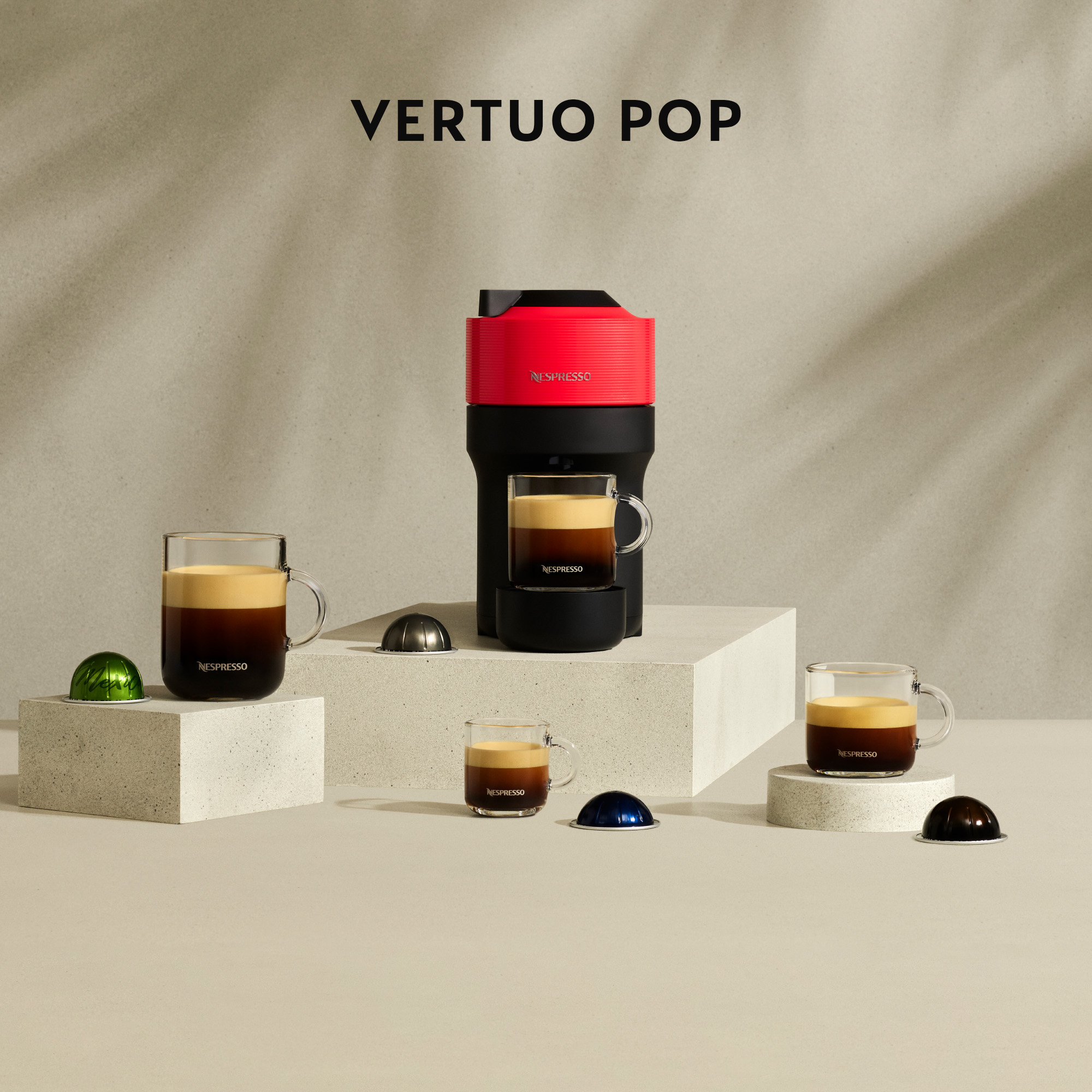 Una máquina Nespresso Vertuo Pop con tazas de café y cápsulas, en un estudio. Máquina roja sobre superficie blanca.