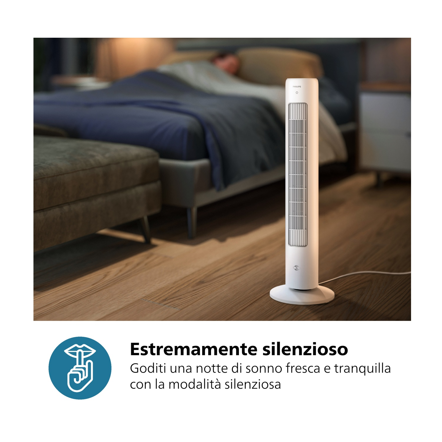 Un ventilatore a torre bianco è su un pavimento in legno in una camera da letto. Sullo sfondo, un letto con lenzuola blu e una persona che dorme.